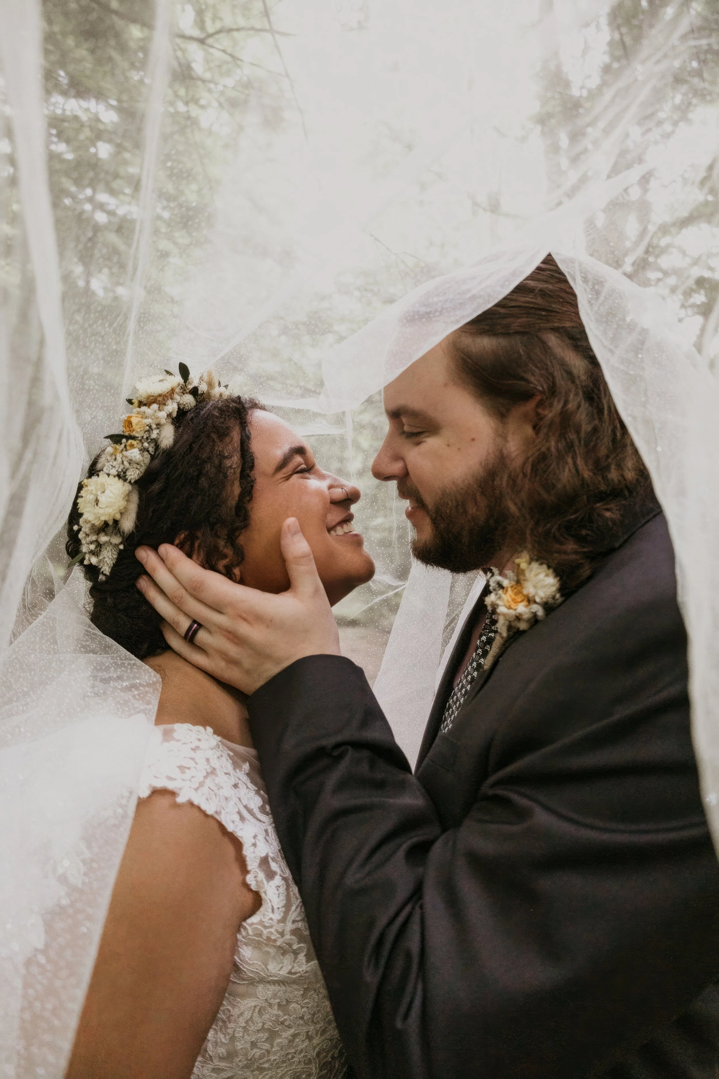 A happy bride and groom on their wedding day under a veil, smiling and holding each other's faces, in a forested outdoor setting.