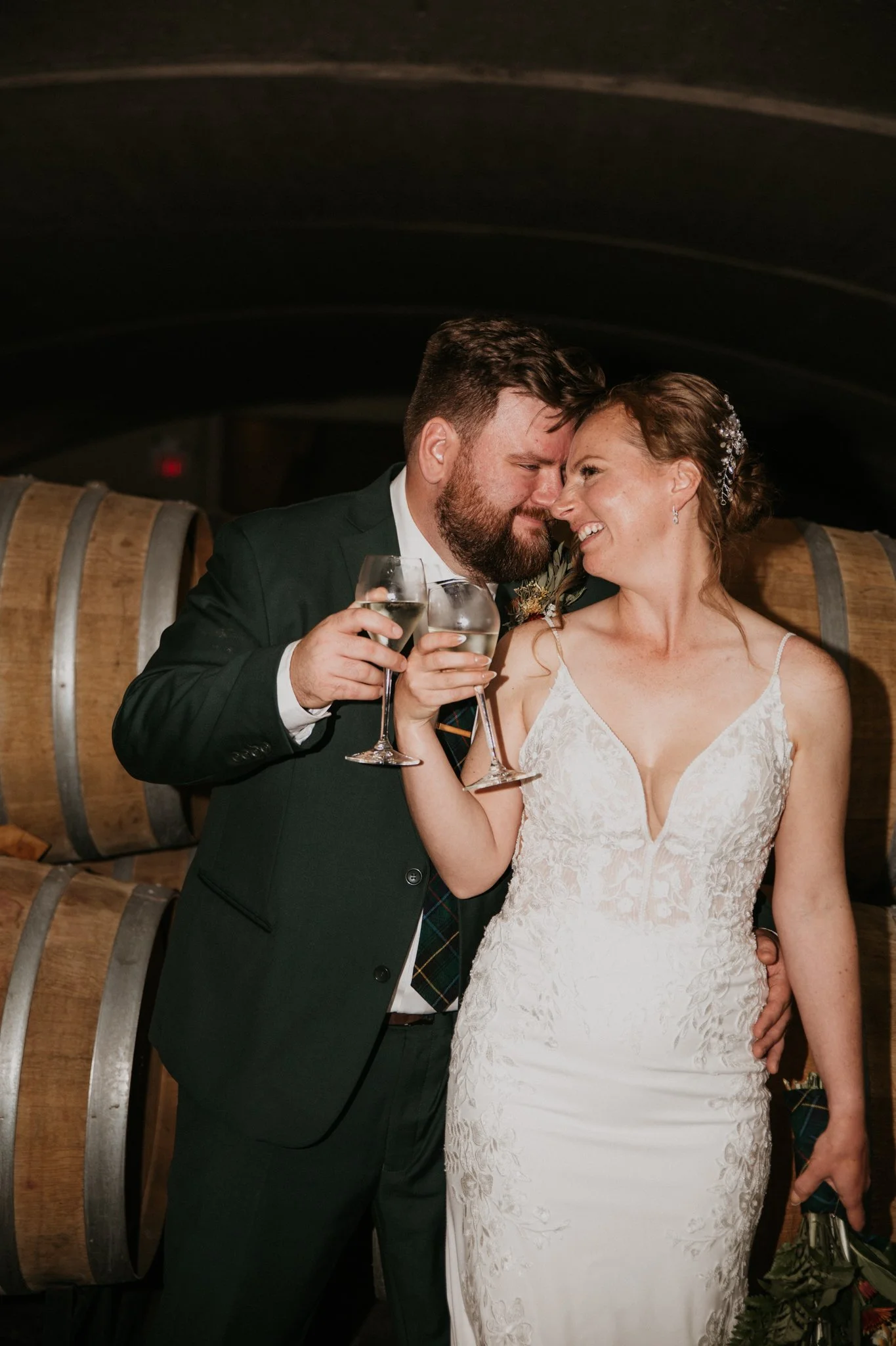 A bride and groom celebrating with wine in a cellar, smiling and touching foreheads.