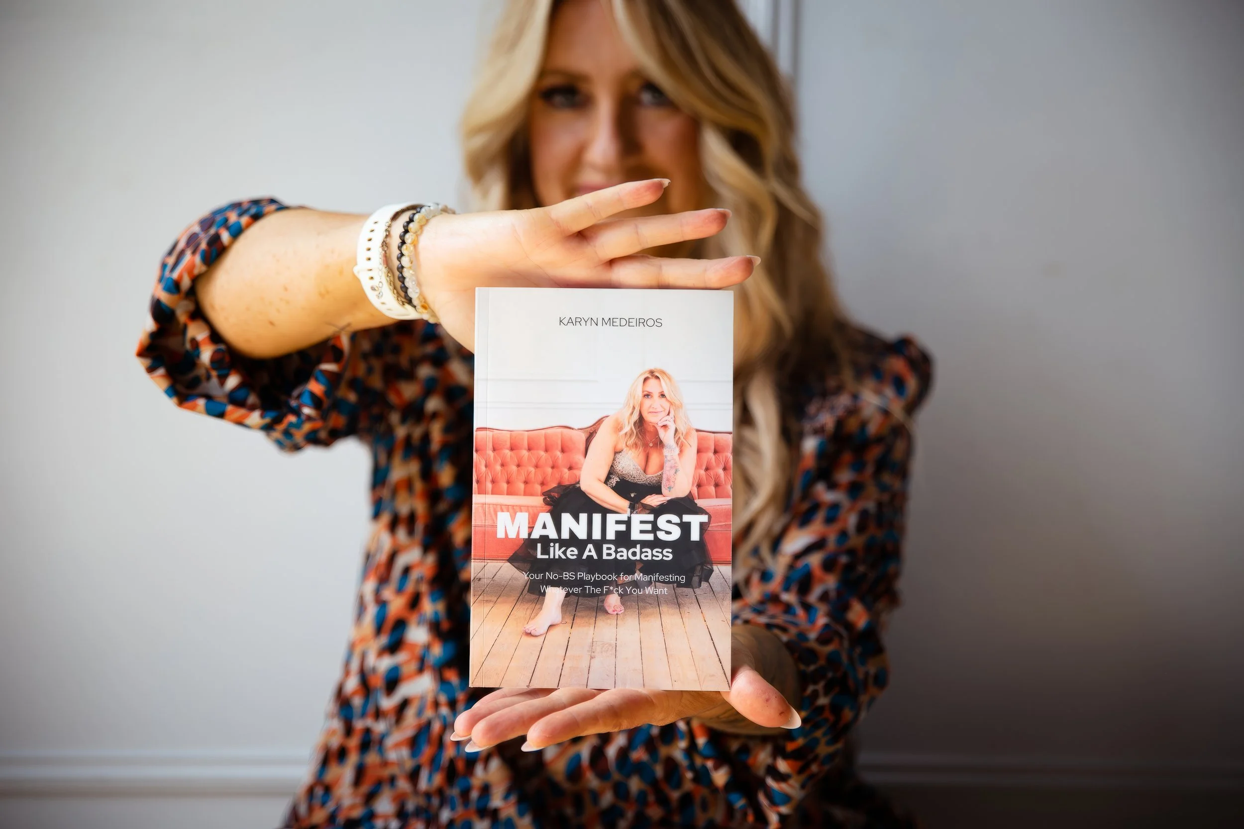 Woman holding a book titled 'Manifest Like a Badass' by Karyn Medeiros, with a photo of a woman sitting on a pink couch on the cover.