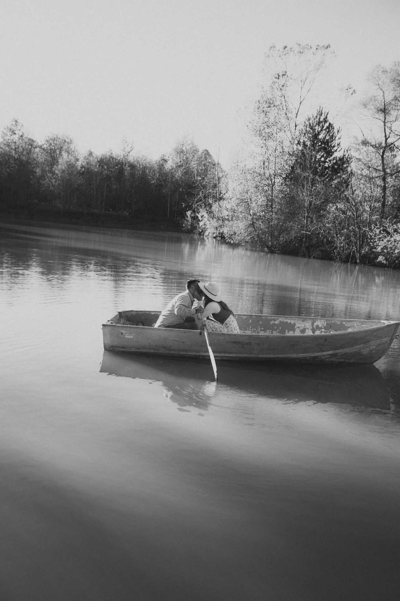 A black and white photo of a couple in a small boat on a calm river, with trees in the background. The woman is wearing a hat and leaning towards the man, who is facing her.