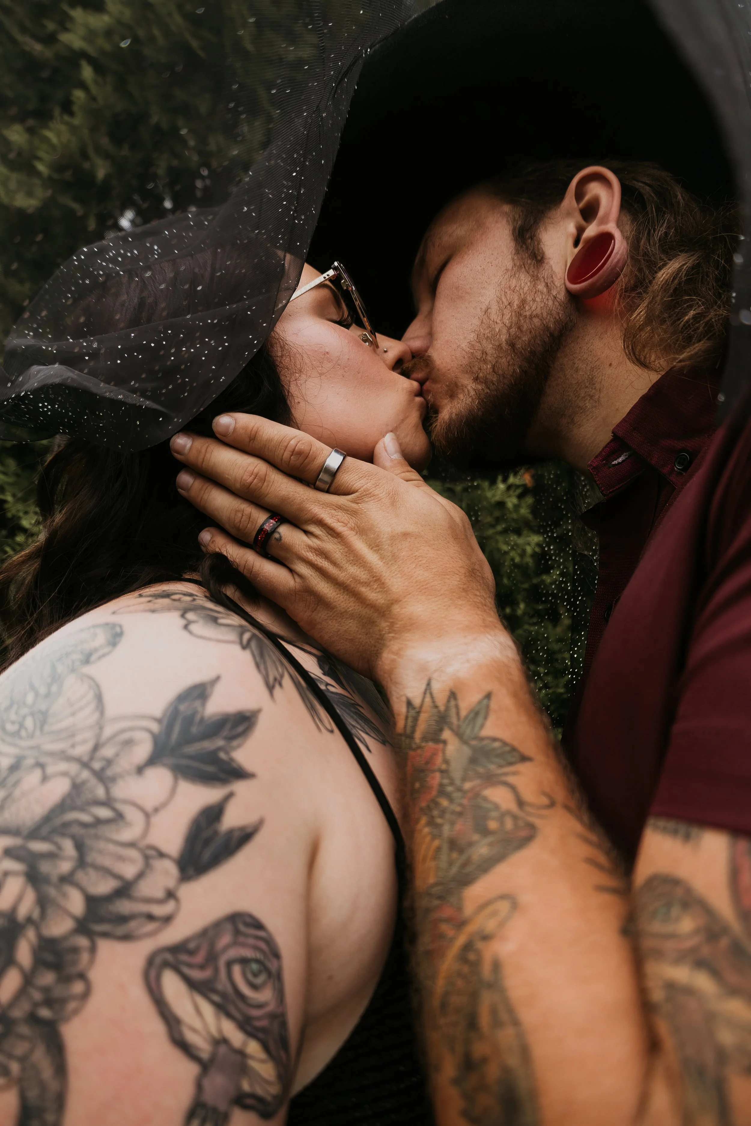 A close-up of a couple kissing outdoors, with tattoos visible on the woman's arm and shoulder, and the man wearing a hat.