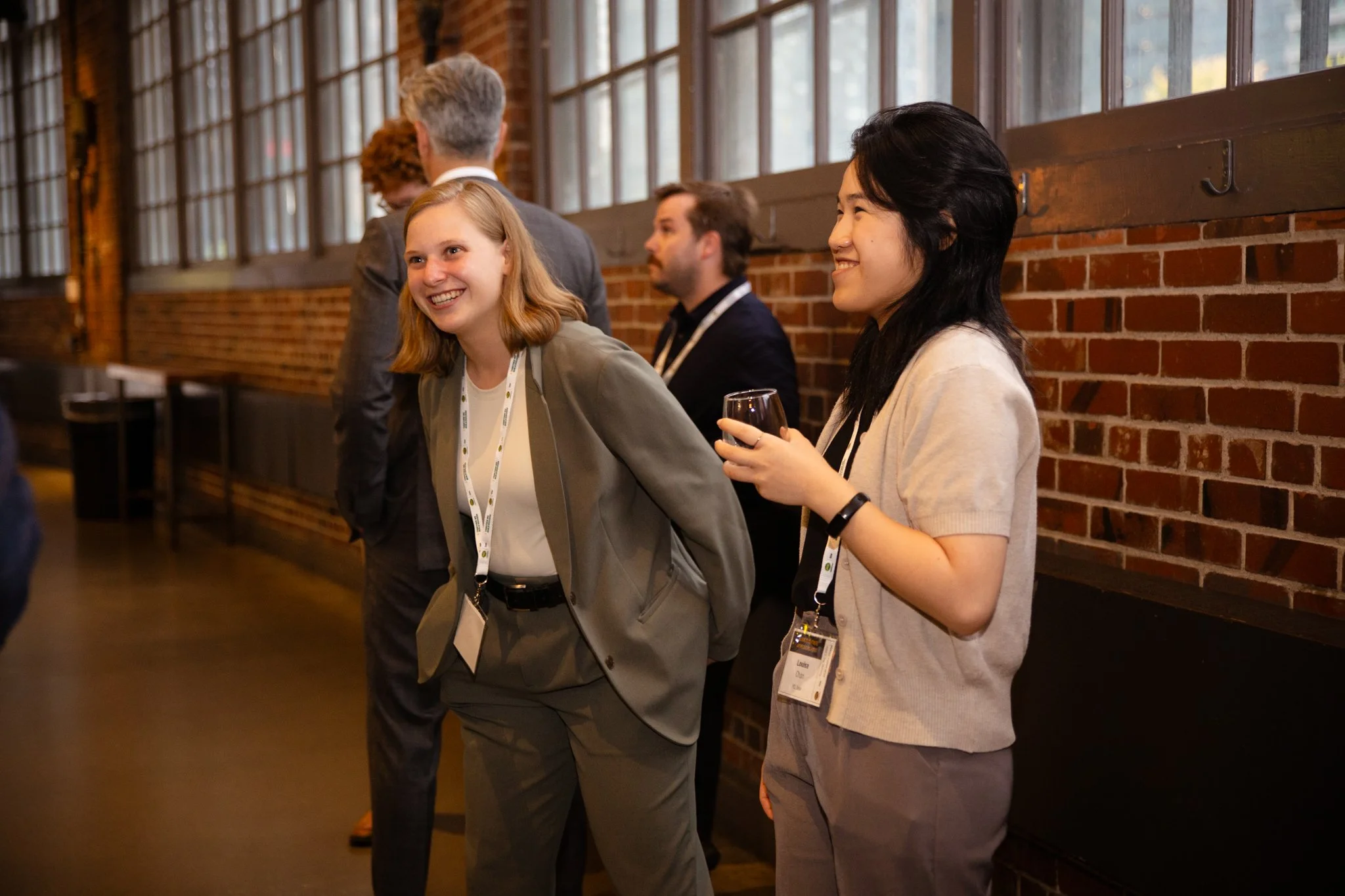 Two women engaged in conversation at a professional networking event, with other people talking in the background against a brick wall.