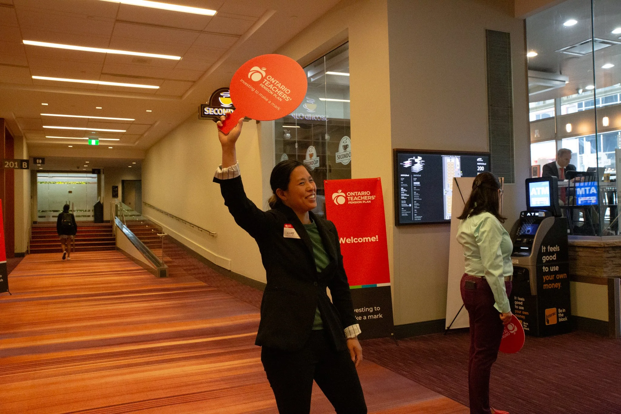 A woman smiling and holding up a red sign that reads 'Ontario Teachers' Pension Plan' inside a lobby or corridor, with a digital display board and a person at a nearby ATM in the background.