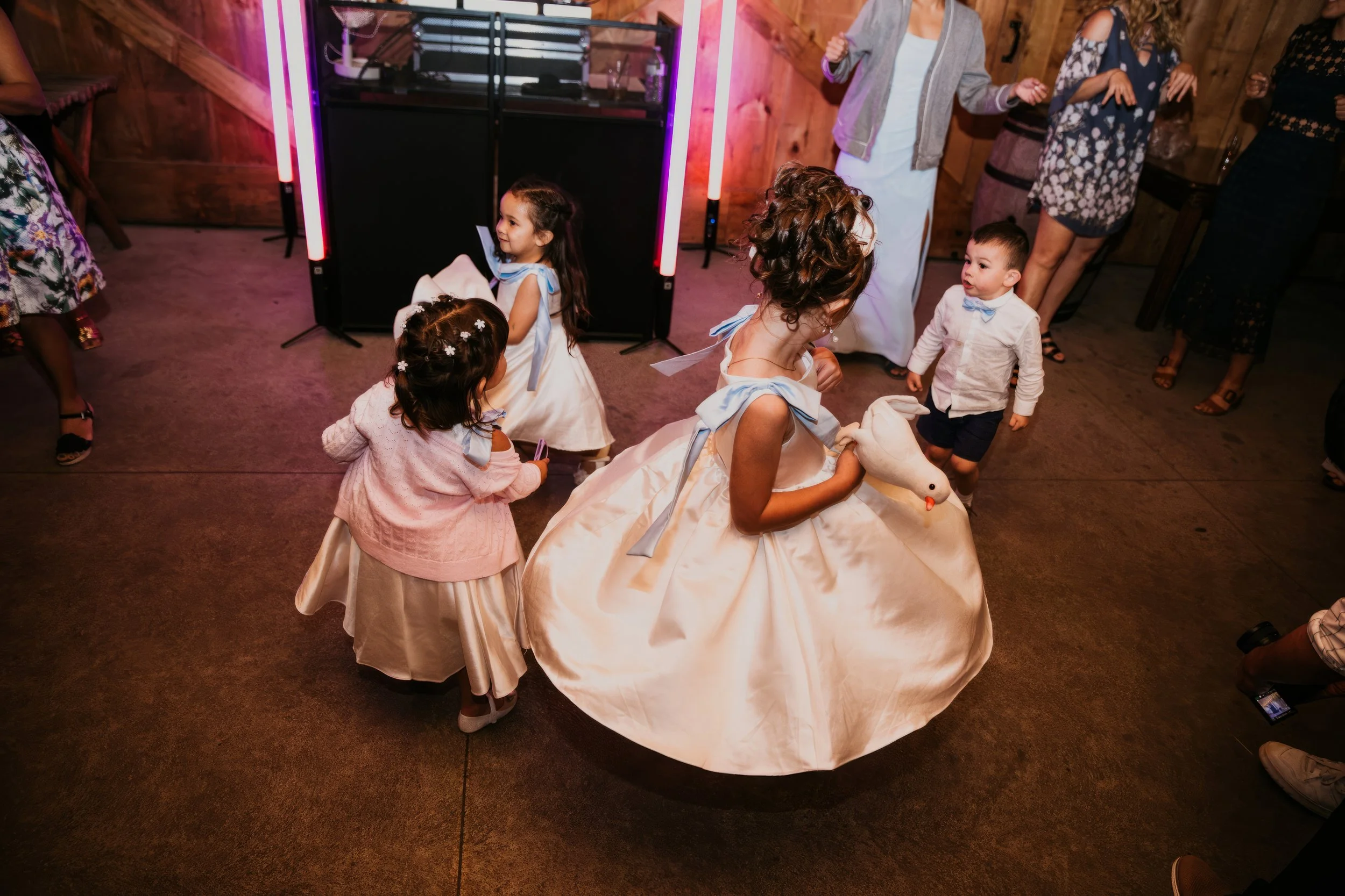 Children dancing at a party, with a woman in a white gown holding a stuffed swan, and others wearing dresses with blue ribbons, in a rustic wooden venue.