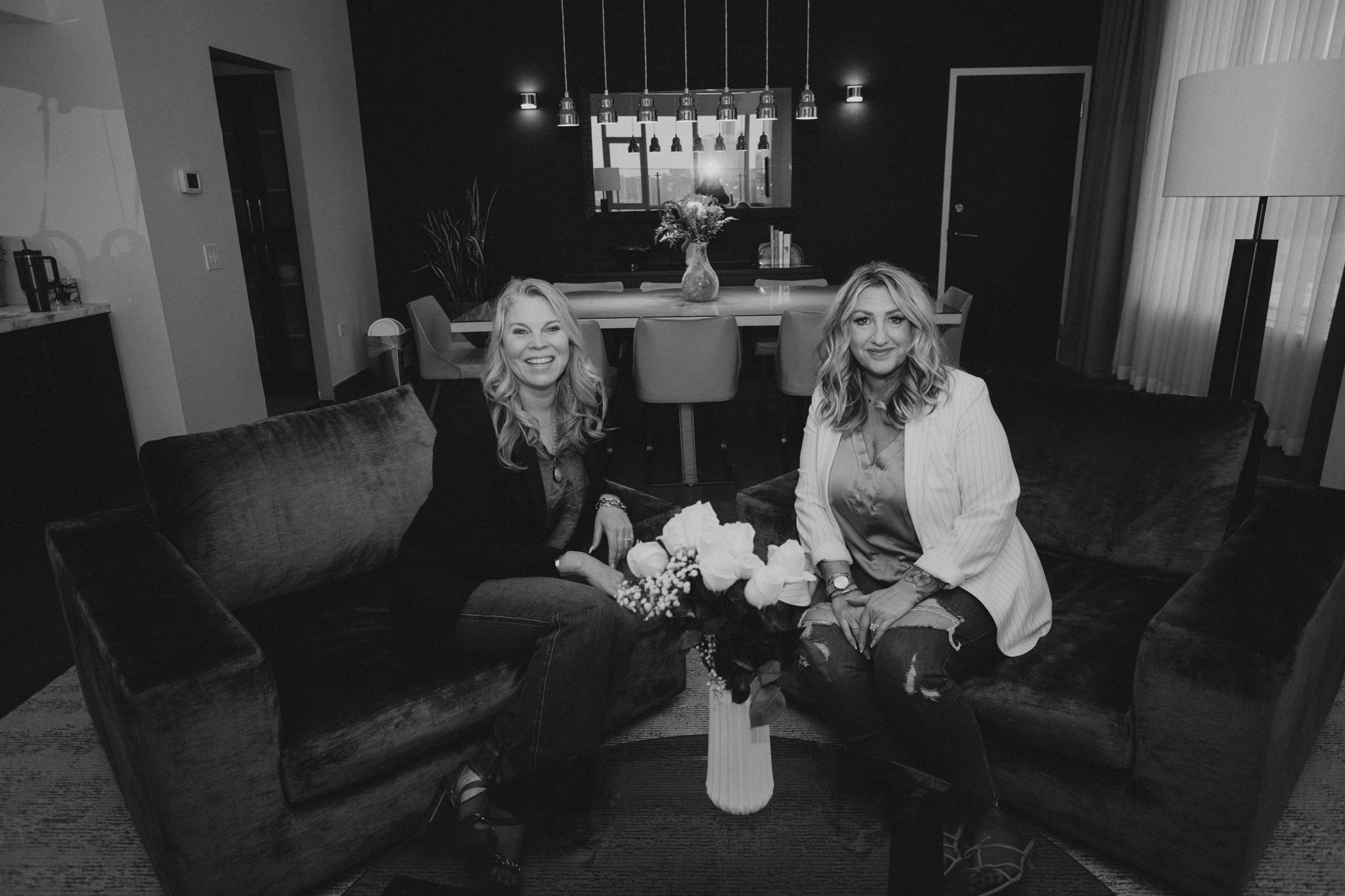 Two women sitting on a dark sofa with a flower arrangement between them, smiling at the camera in a modern living room with a dining area in the background.