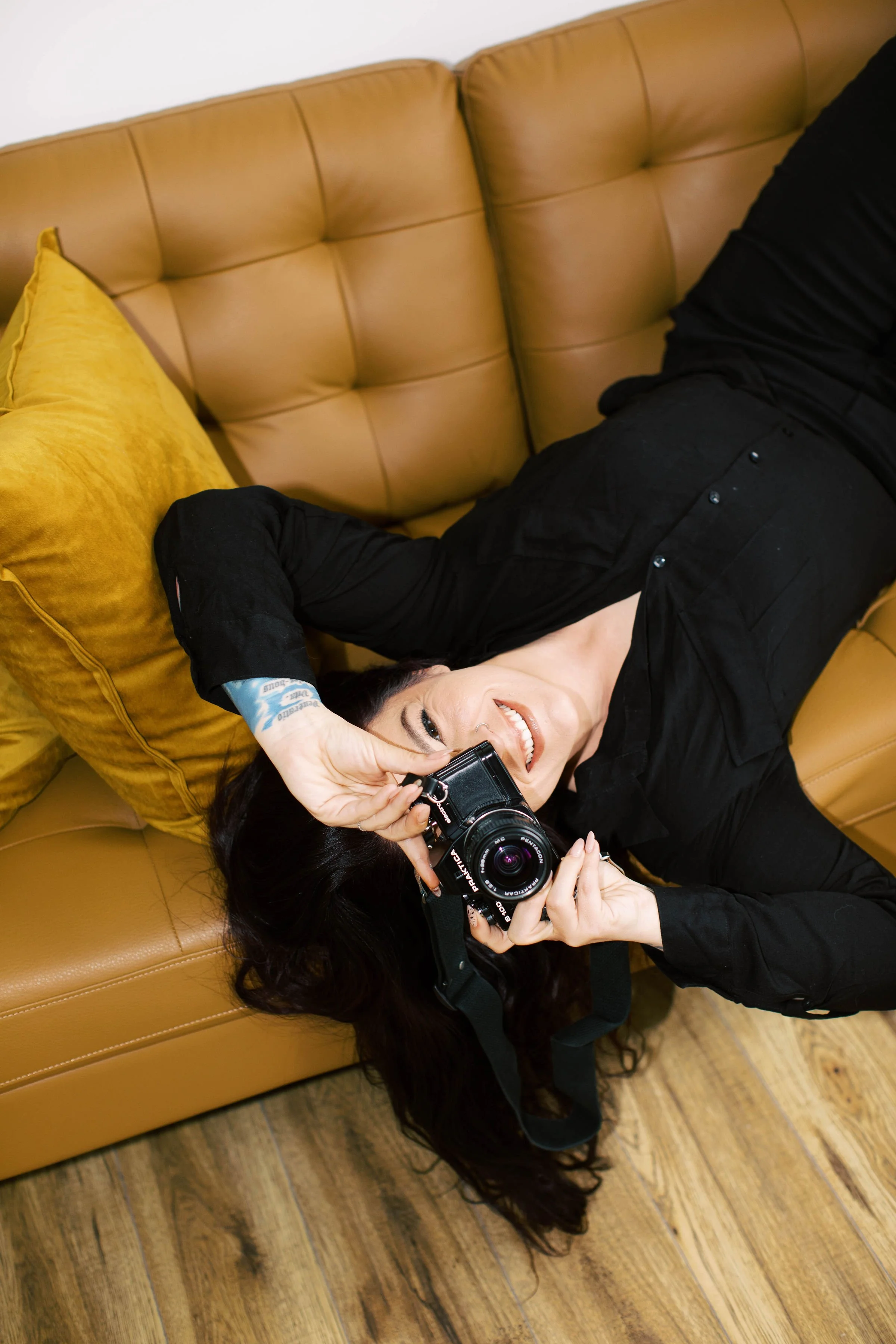 A woman with black hair lies on a tan leather couch, smiling and holding a camera to her eye, with a yellow pillow behind her head.