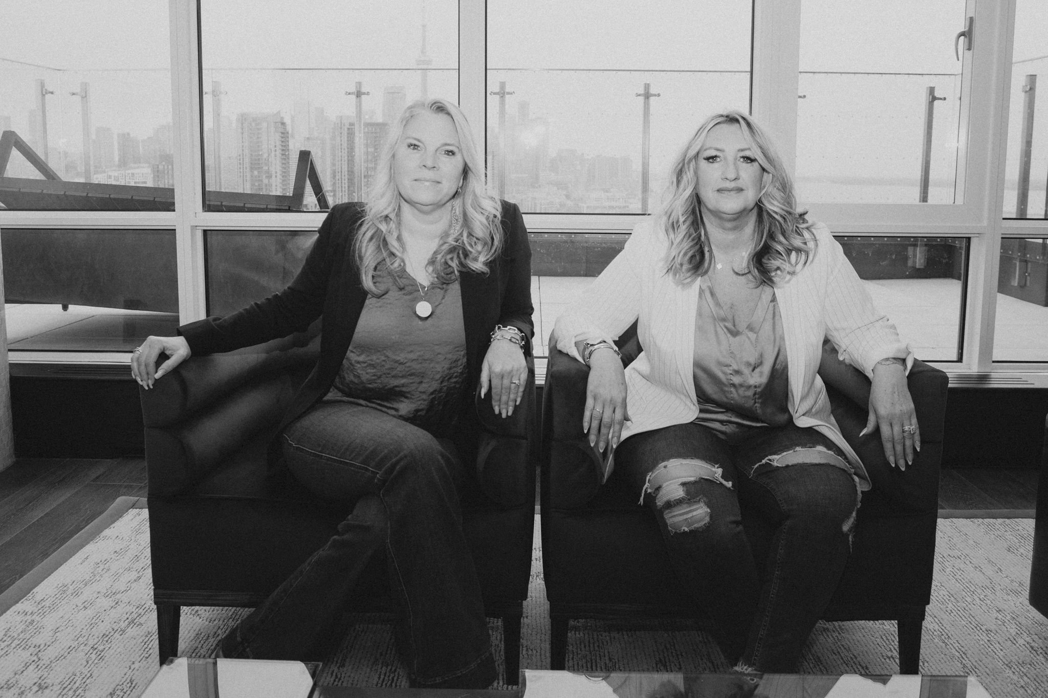 Two women sitting on black sofas in a bright room with large windows showing a cityscape in the background.