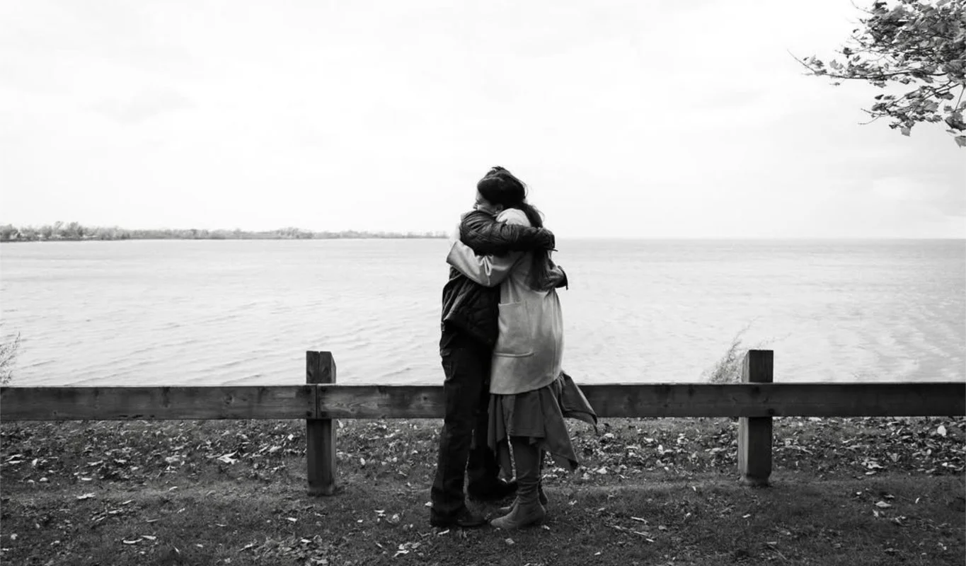 A couple hugging near a lake with a wooden barrier in the foreground and trees on the right.