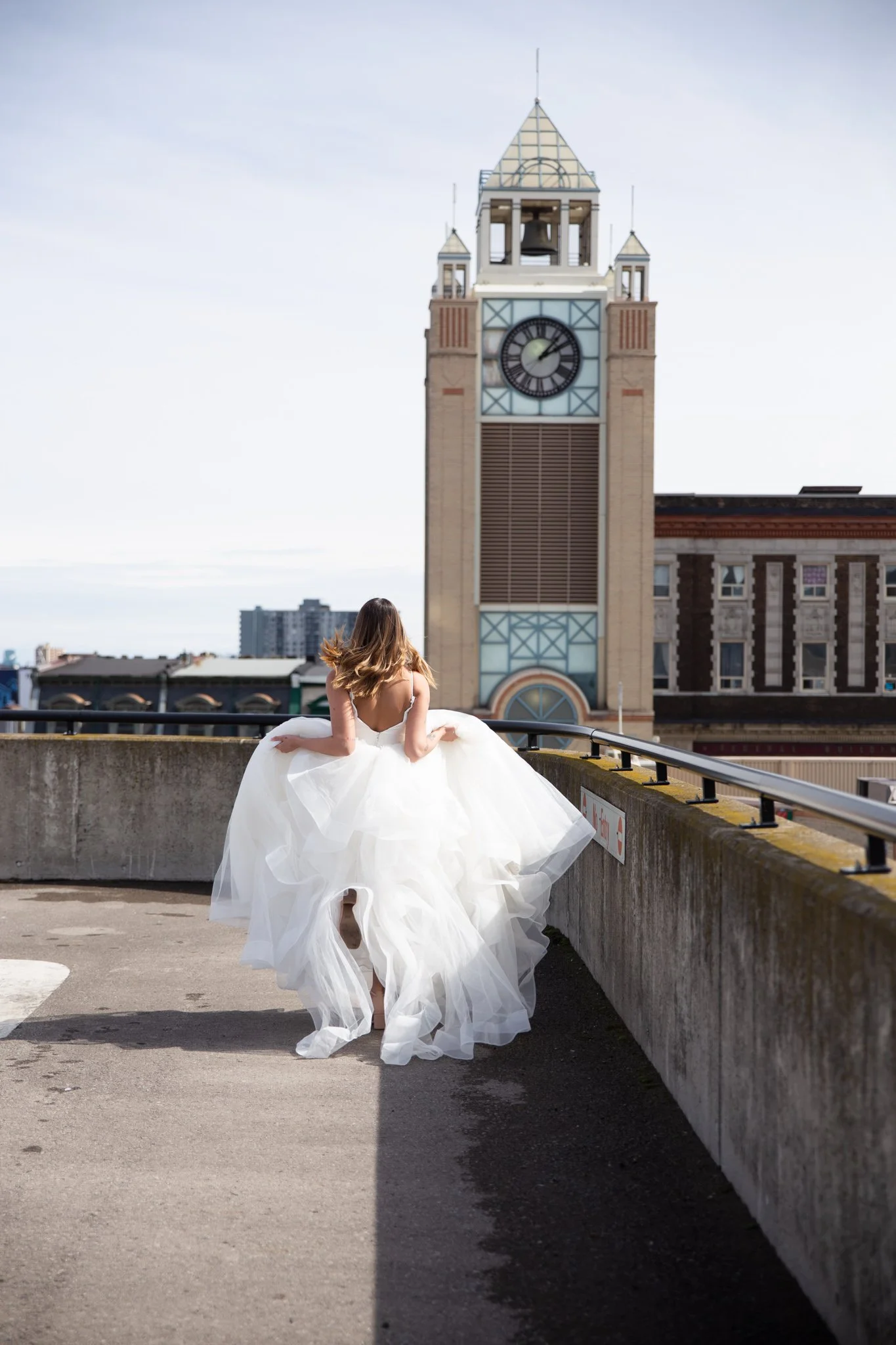 A woman in a white wedding dress walking on a parking garage rooftop, with a large clock tower in the background.