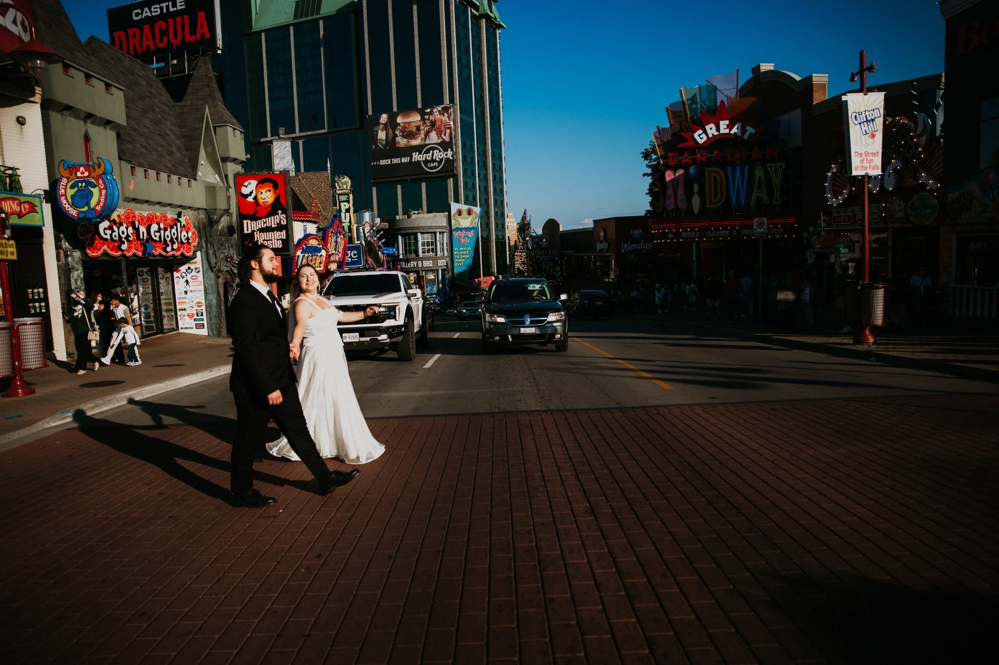A bride and groom walk across a brick street in a lively urban area with various storefronts, bright signs, and parked cars on a sunny day. The bride wears a white gown, and the groom is in a black suit.