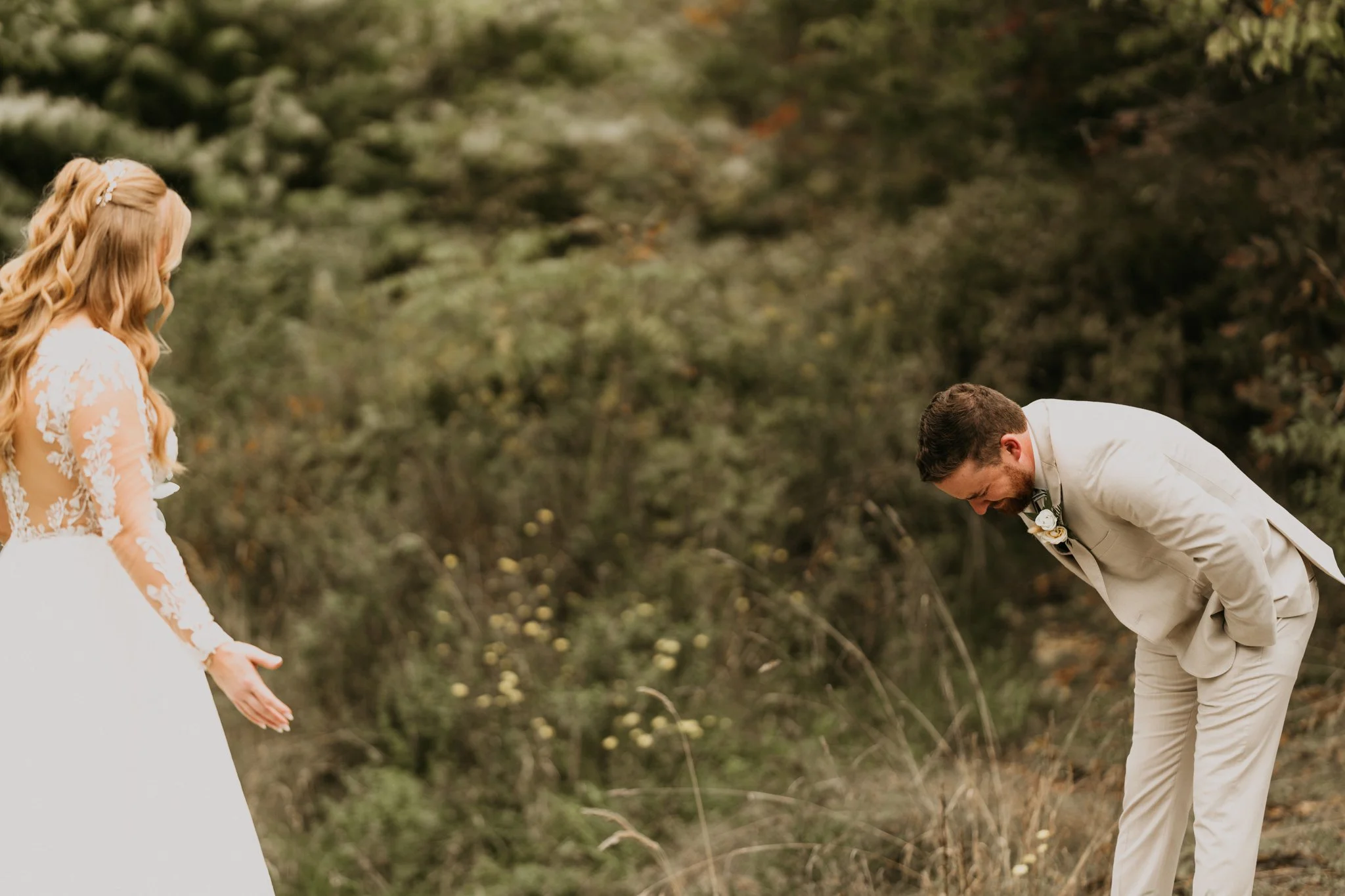 A bride in a white wedding gown with lace details appears surprised or emotional, looking at a groom in a light-colored suit with a boutonniere, bowing or bending forward in front of her in an outdoor setting with trees and greenery.