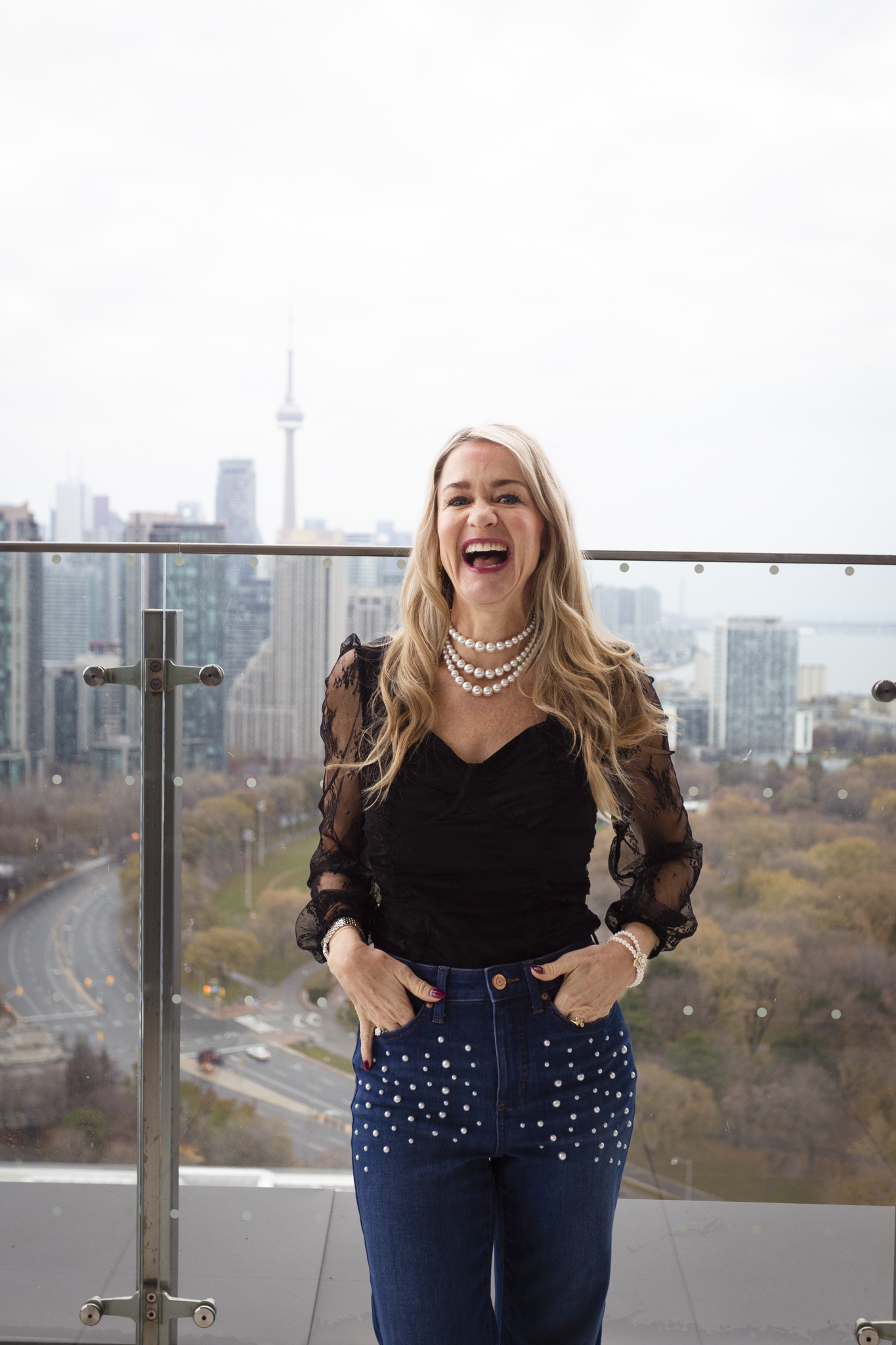 A woman laughing on a rooftop balcony with a cityscape in the background, wearing a black lace top and pearl jewelry.
