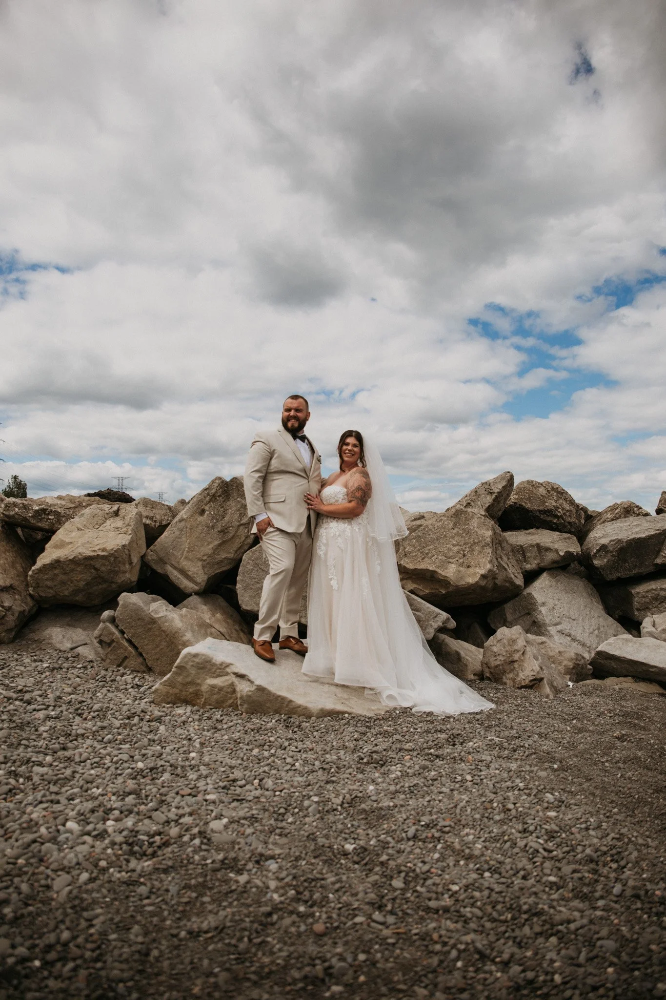 A bride and groom on a large rock, smiling, with a cloudy sky backdrop.