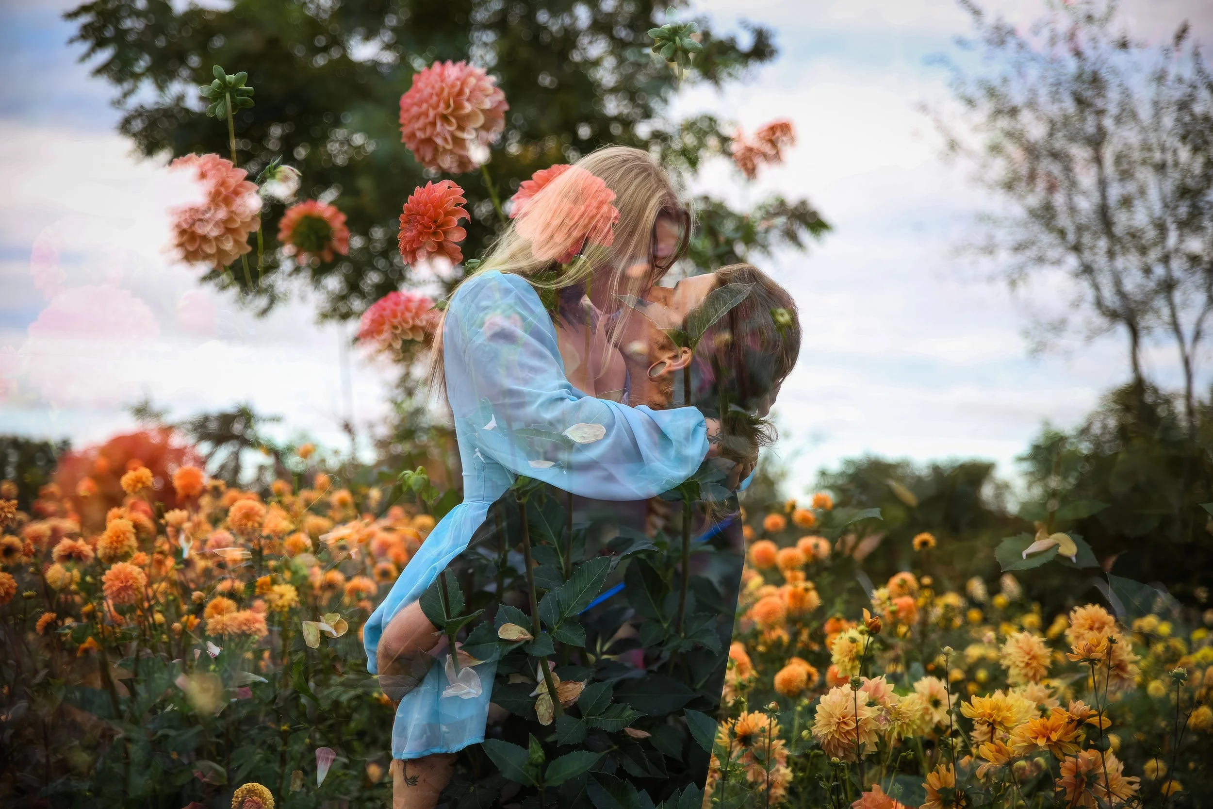 A couple embracing in a garden filled with orange and yellow flowers, with a partly cloudy sky and trees in the background.
