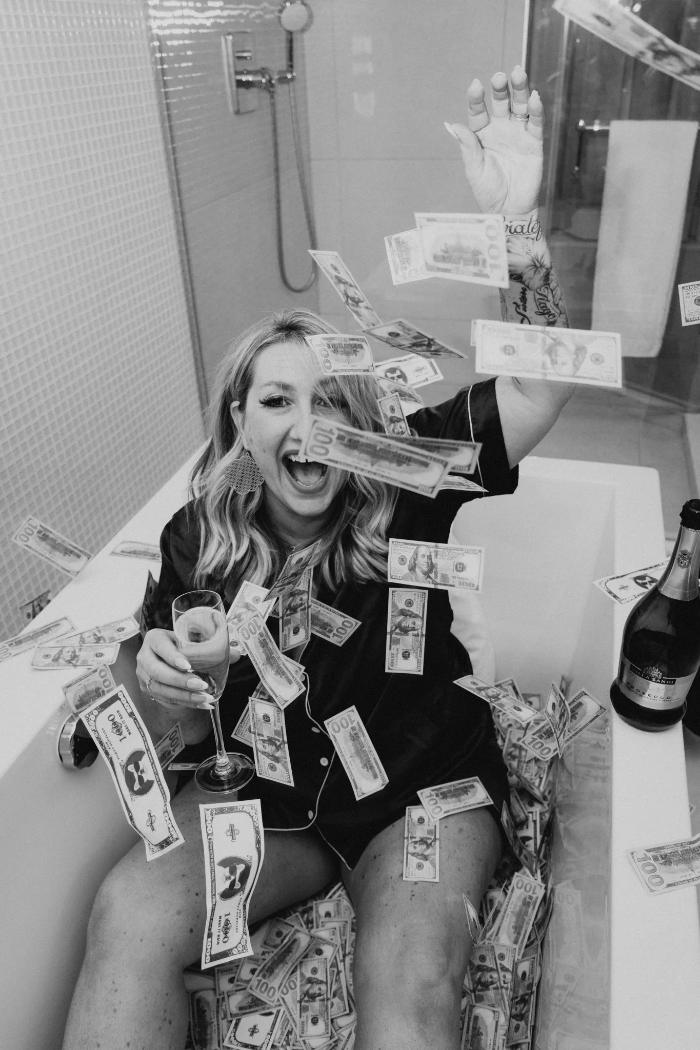 A woman in a bathtub with money falling around her, holding a glass of champagne and smiling joyfully.