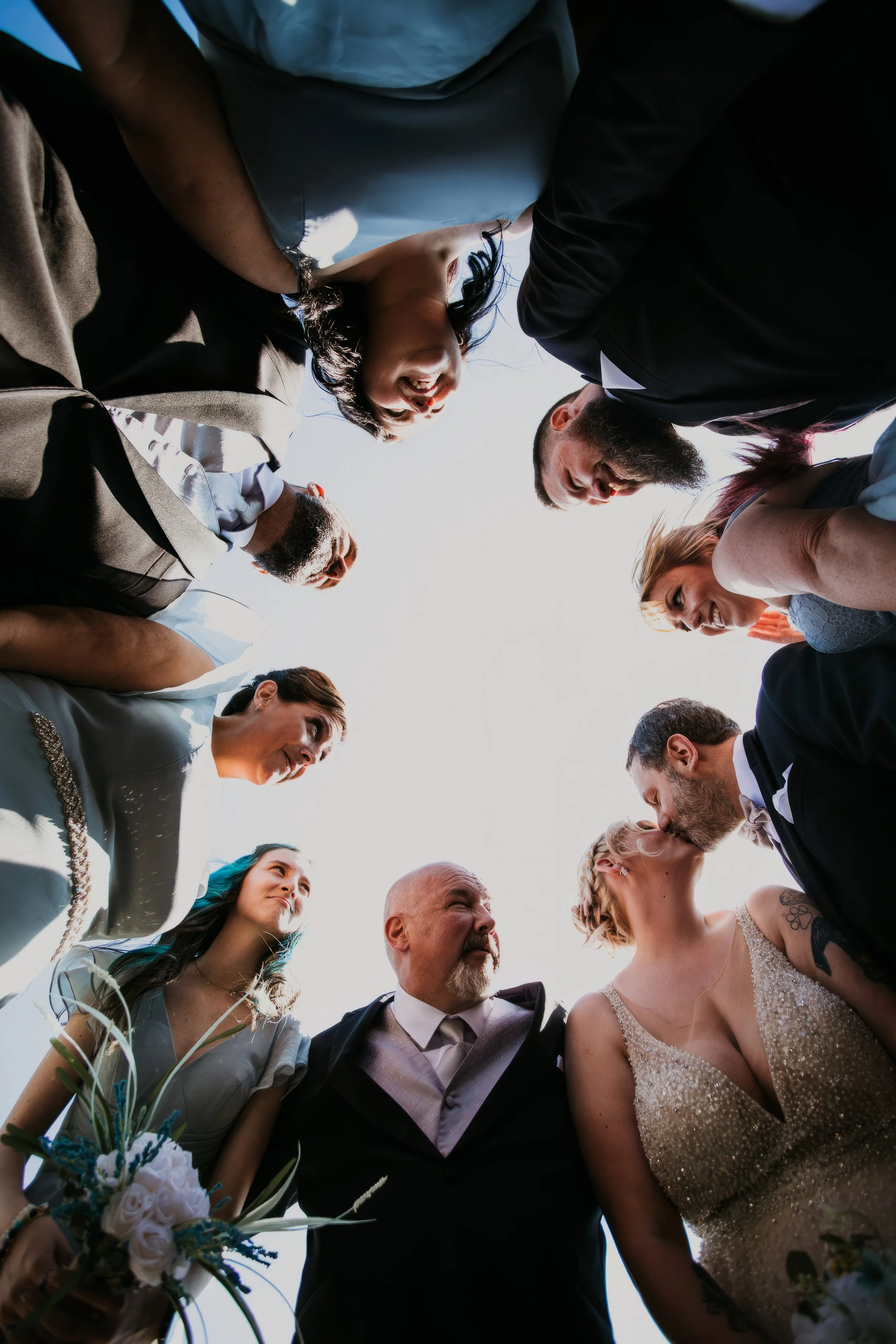 A group of people, including a bride and groom, gathered in a circle at a wedding, looking down at the camera positioned below.