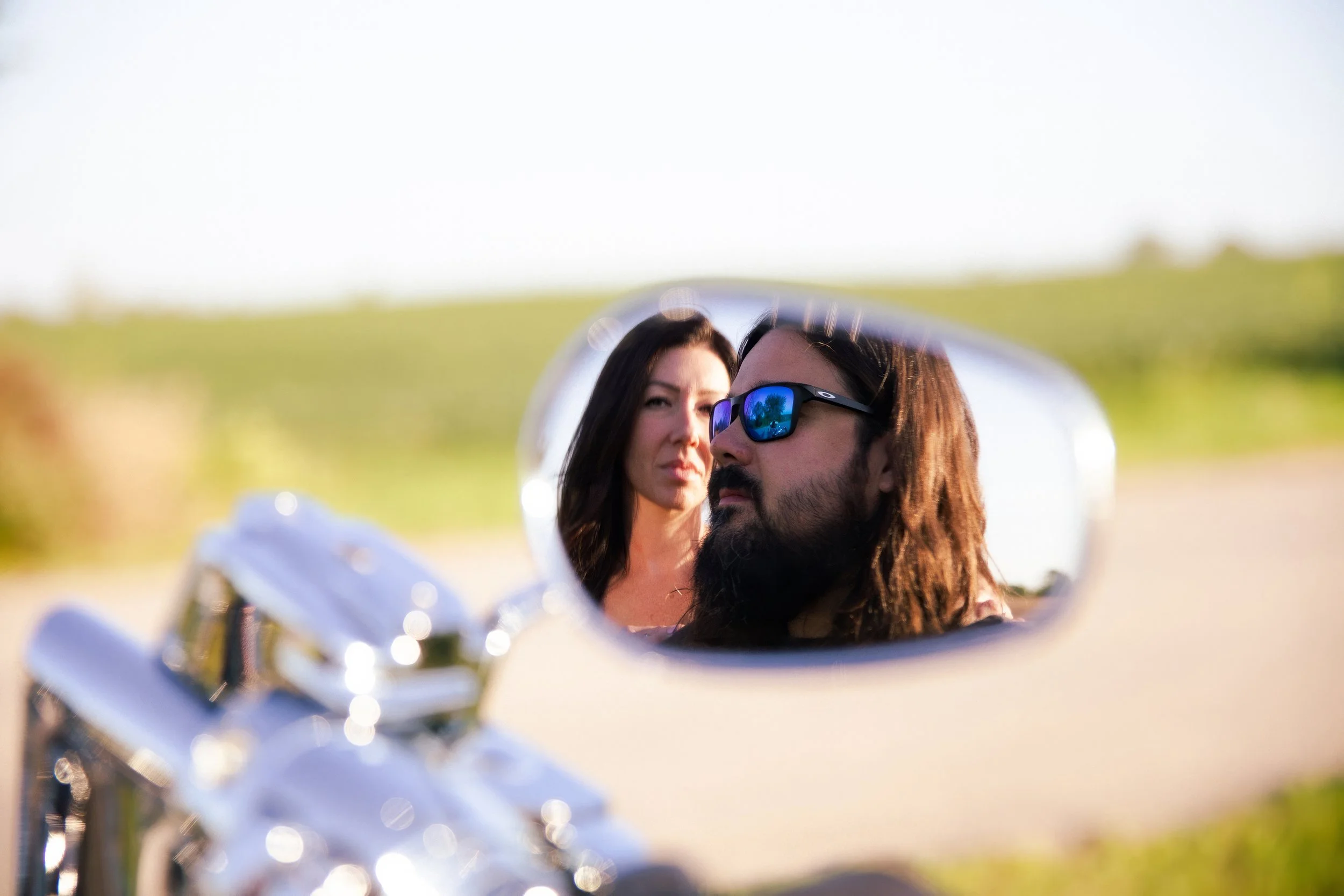 Reflection of a man with long hair and sunglasses and a woman with dark hair in a motorcycle mirror against a blurred outdoor background.