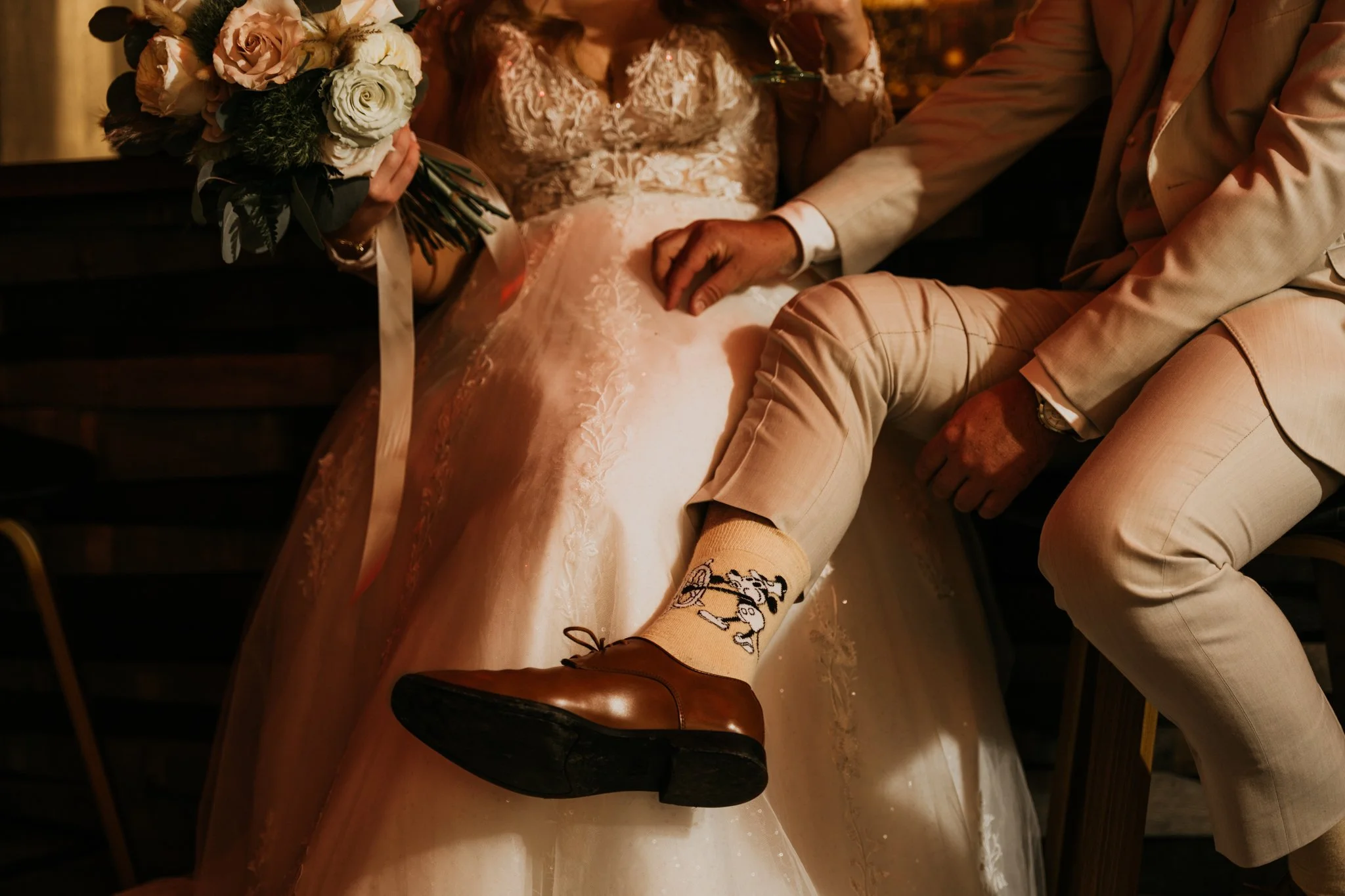 A bride holding a bouquet of pink and white flowers with greenery, sitting next to a groom dressed in a beige suit, with the groom's leg crossed over the bride's lap, showing a Snoopy character on his sock and polished brown shoes.