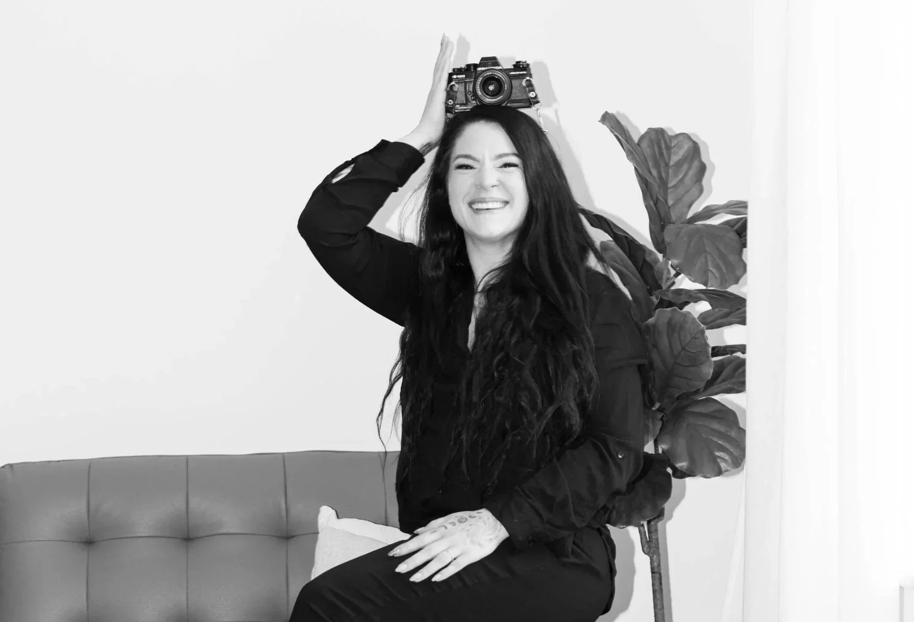 Black and white photo of a woman with long dark hair smiling, holding a camera above her head, sitting on a couch next to a large plant.