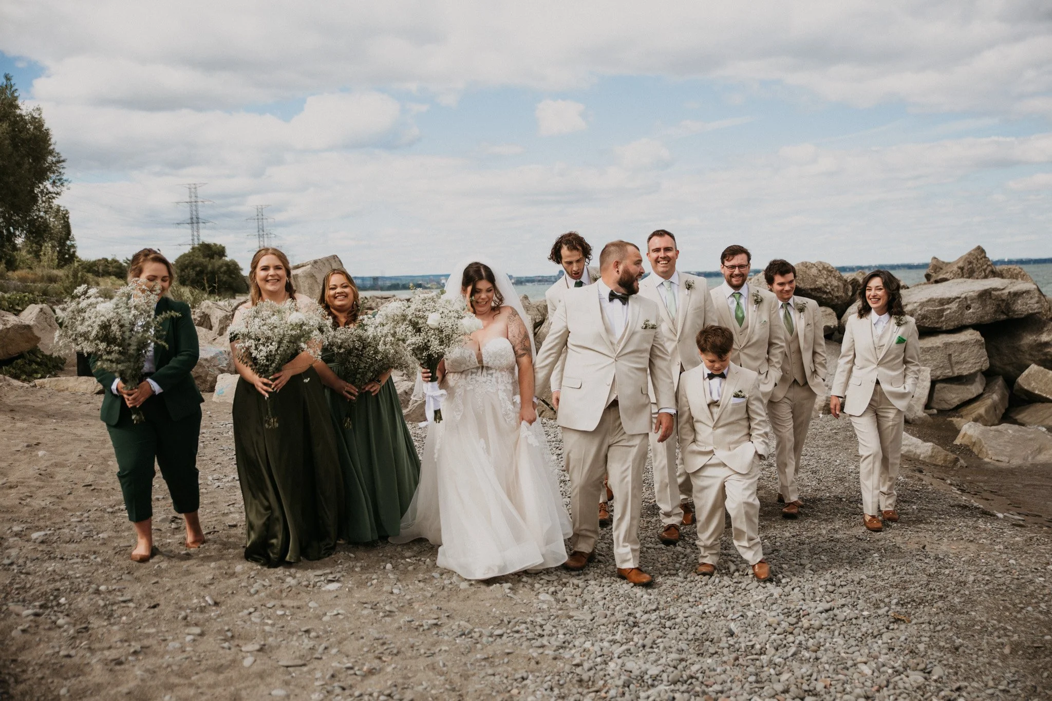 A wedding party walking outdoors on a rocky path near water, with cloudy sky and rocks in the background. The bride in a white gown is accompanied by bridesmaids in dark green dresses, and groomsmen in light suits with ties or bow ties.
