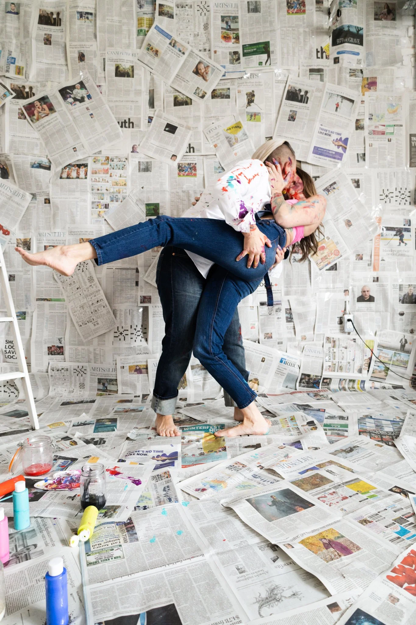 Two people kissing, one lifting the other, in a room covered with scattered newspapers and some paint supplies on the floor.
