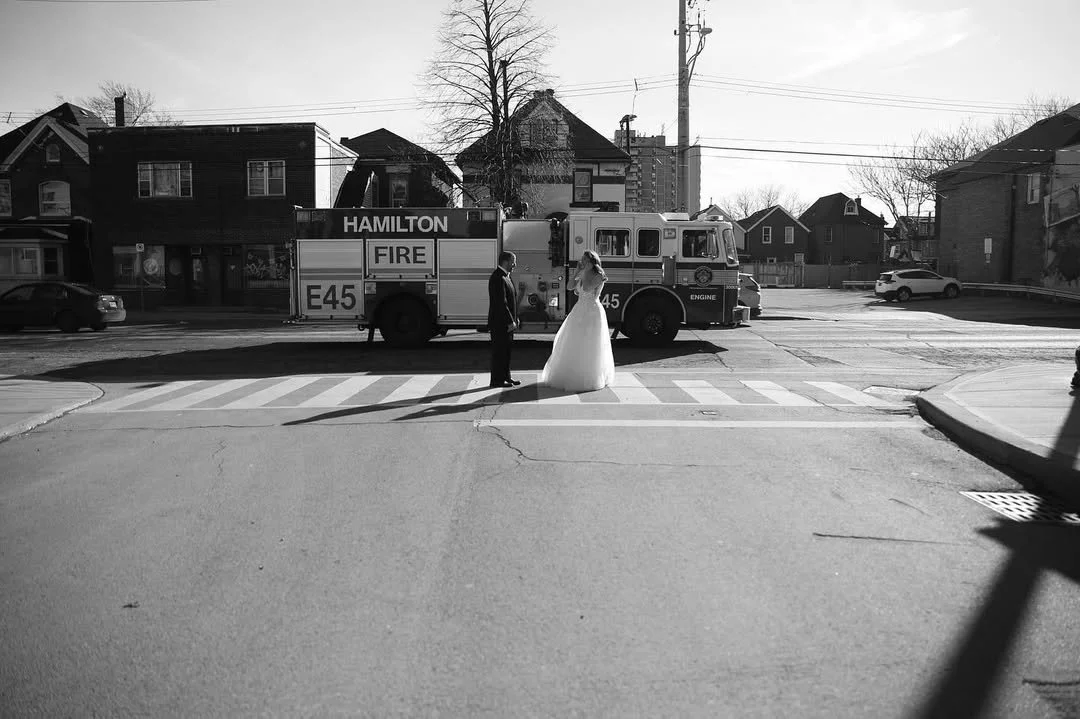 A black and white photo of a bride and groom holding hands on a crosswalk, with a fire truck in the background and houses on either side.