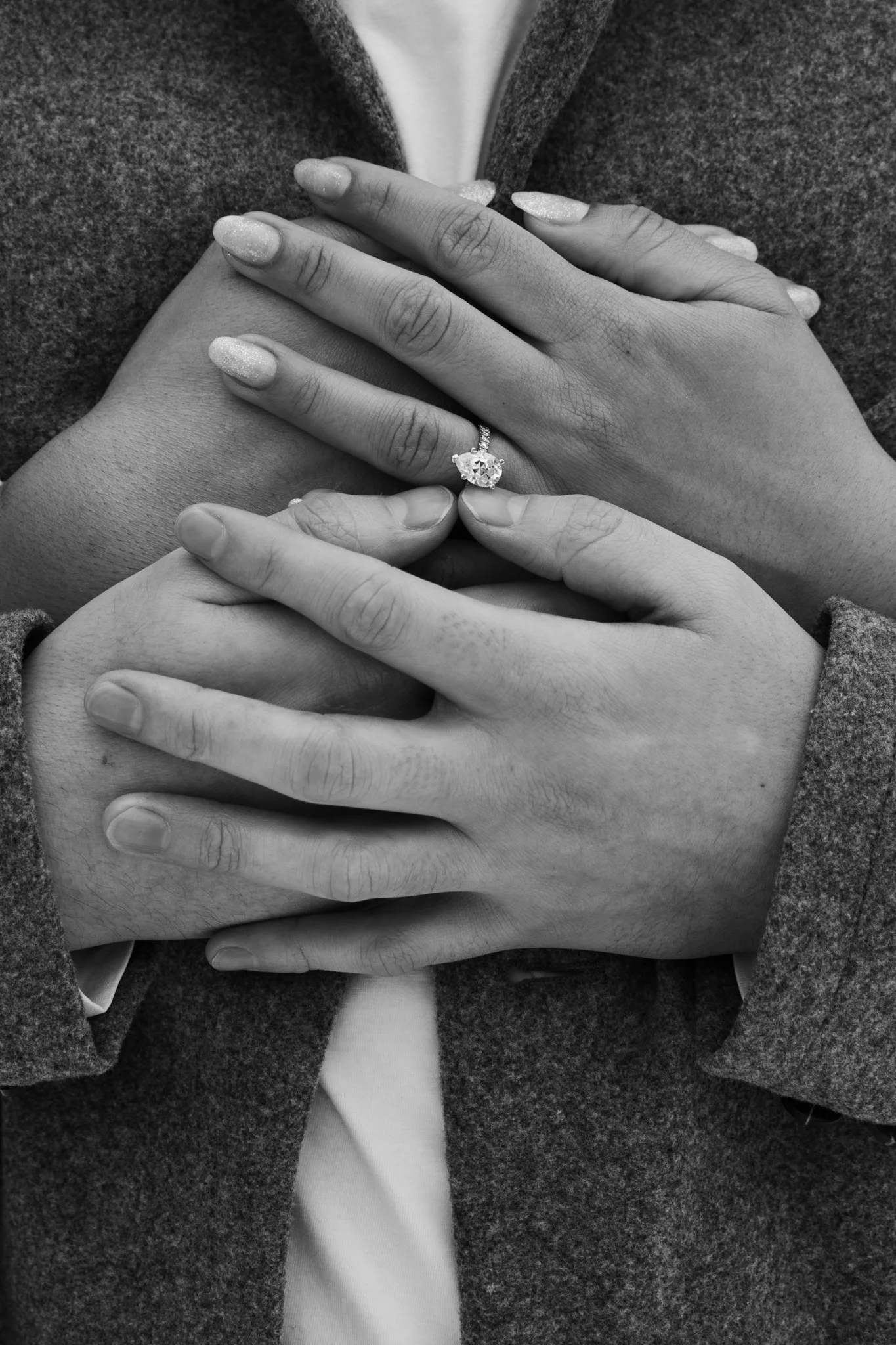 Close-up of a person wearing a dark coat and white shirt, with hands placed on chest, showcasing an engagement ring with a large gemstone on the ring finger of the top hand, and manicured nails.