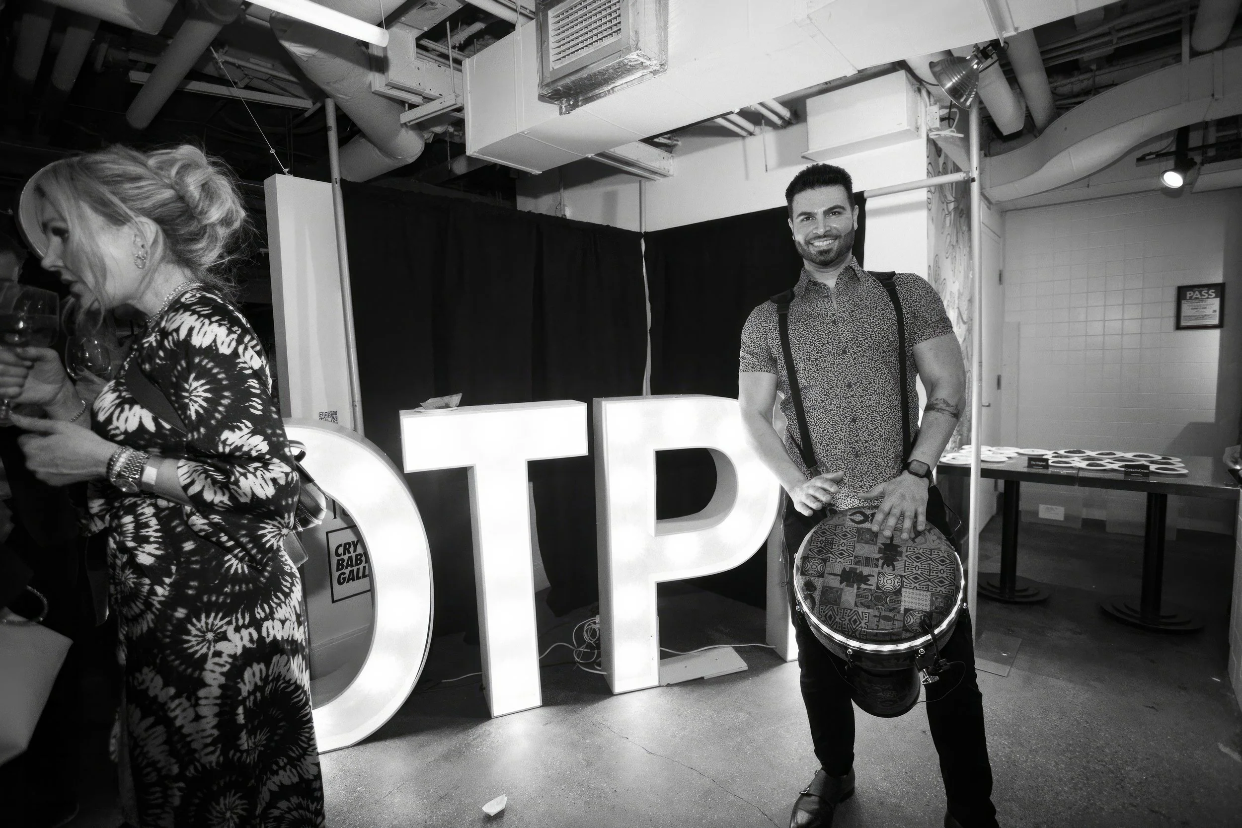 A smiling man standing next to large illuminated letters spelling 'TID' at an indoor event. The man is holding a drum and is wearing a patterned shirt, with a backpack and watch. A woman in a patterned dress is on the left side, holding a drink.