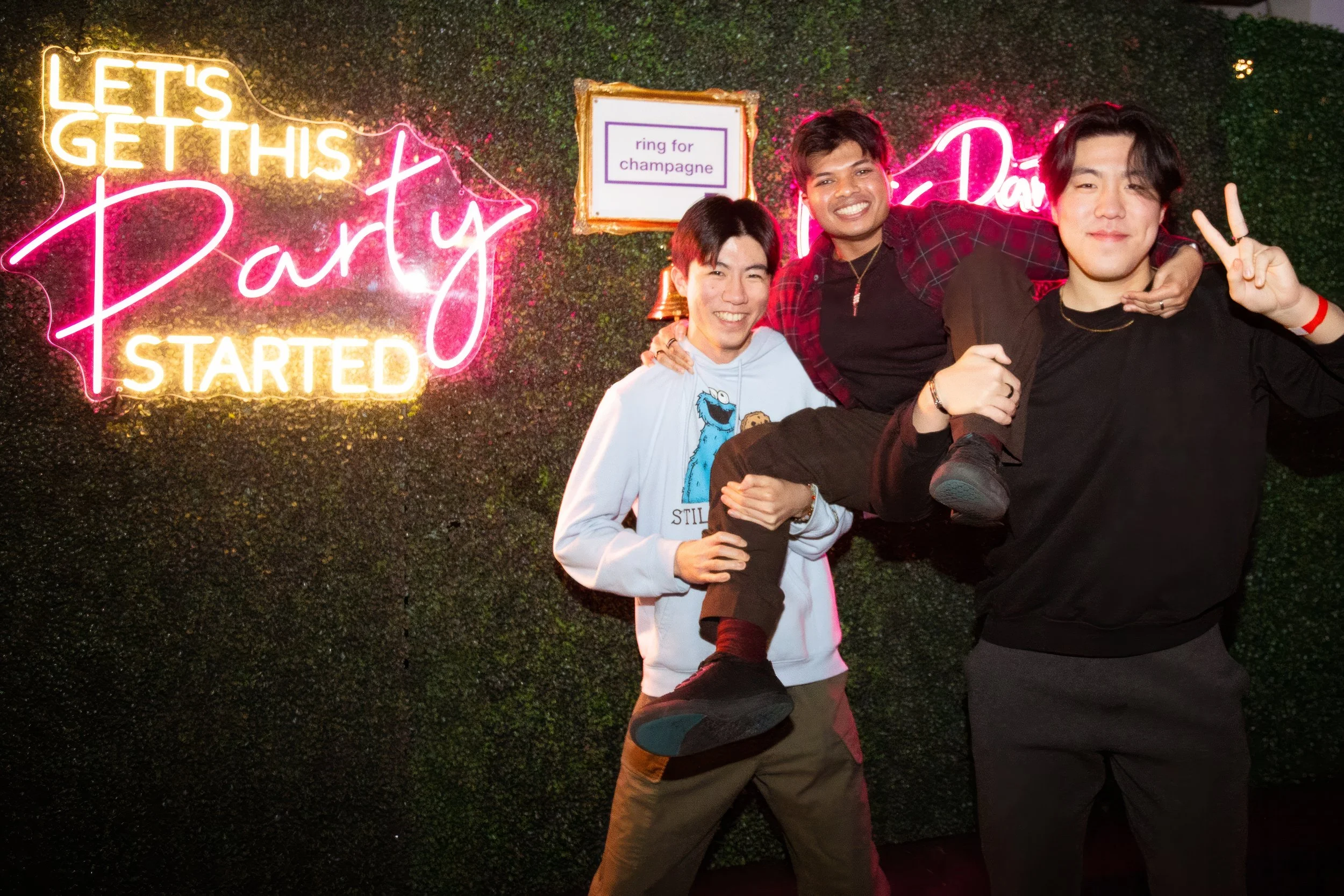 Three men at a party, one giving a piggyback ride, against a wall with neon signs, including 'Let's Get This Party Started' and a small sign that says 'ring for champagne.'