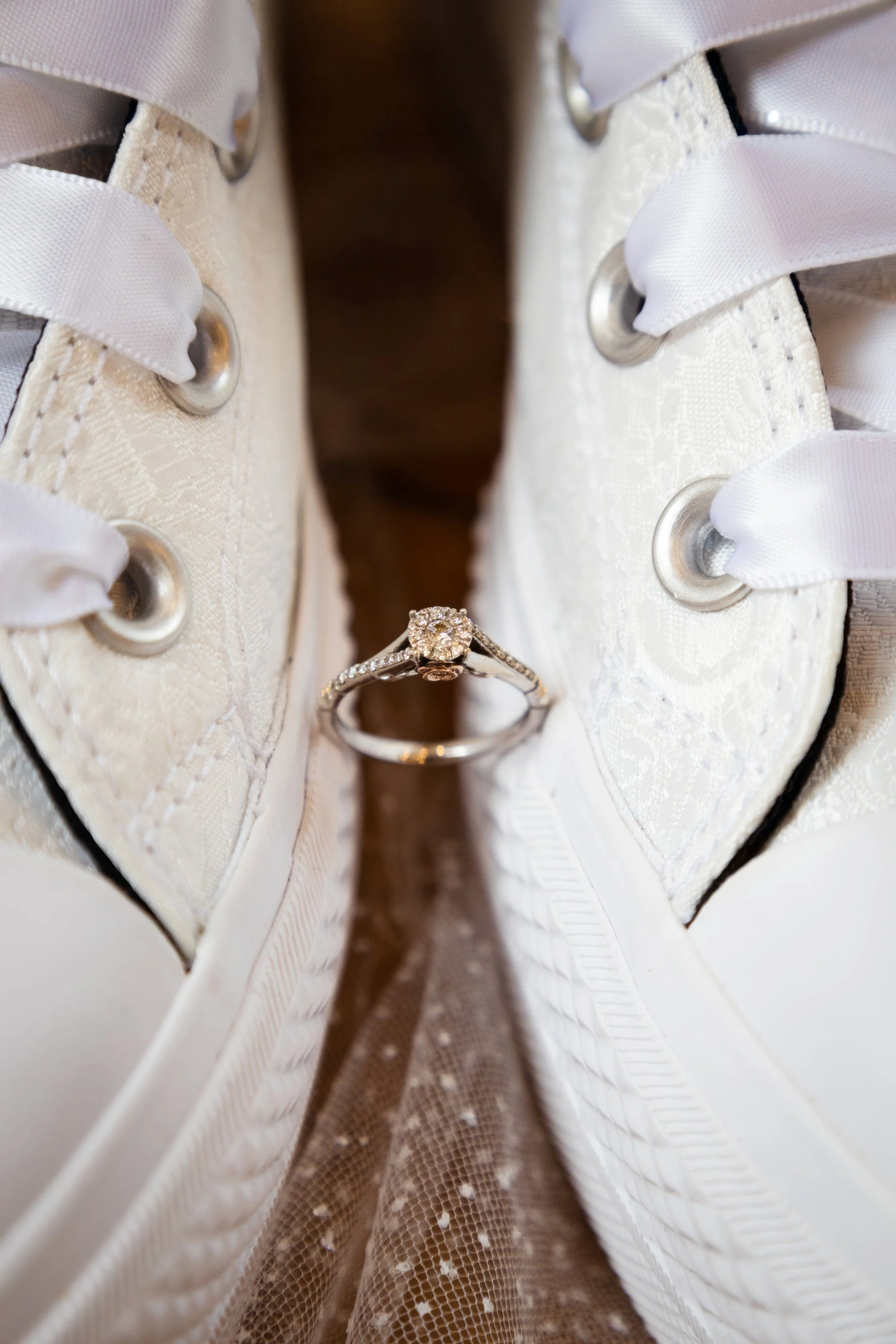 A close-up of white wedding shoes with satin ribbons and eyelet laces, with a diamond engagement ring and wedding band placed between them.