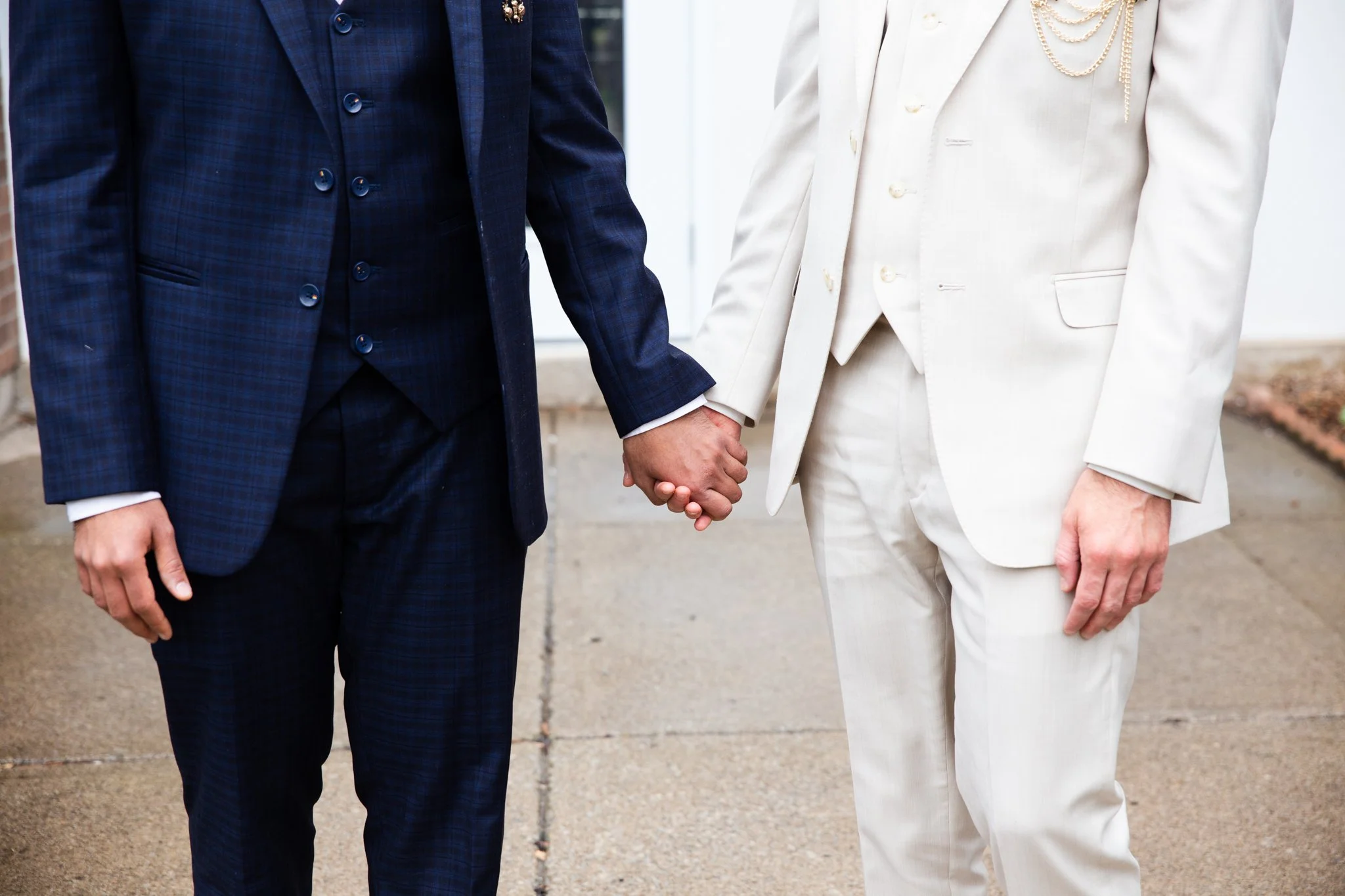 A same-sex couple holding hands dressed in formal suits, standing outdoors.