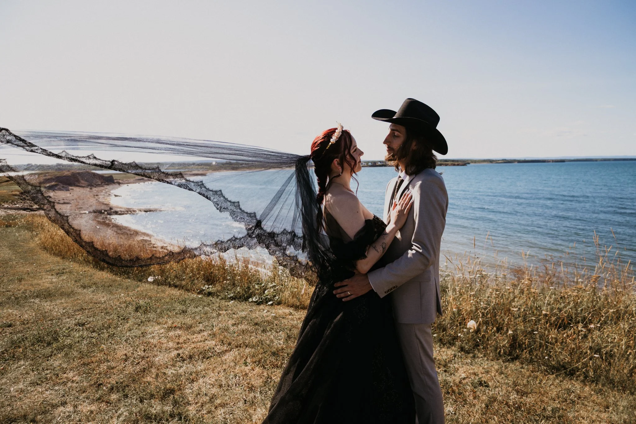 A couple dressed as a bride and groom standing close together on a grassy area near water, with the bride wearing a black dress and lace veil, and the groom wearing a light-colored suit and cowboy hat, holding each other as they look into each other'