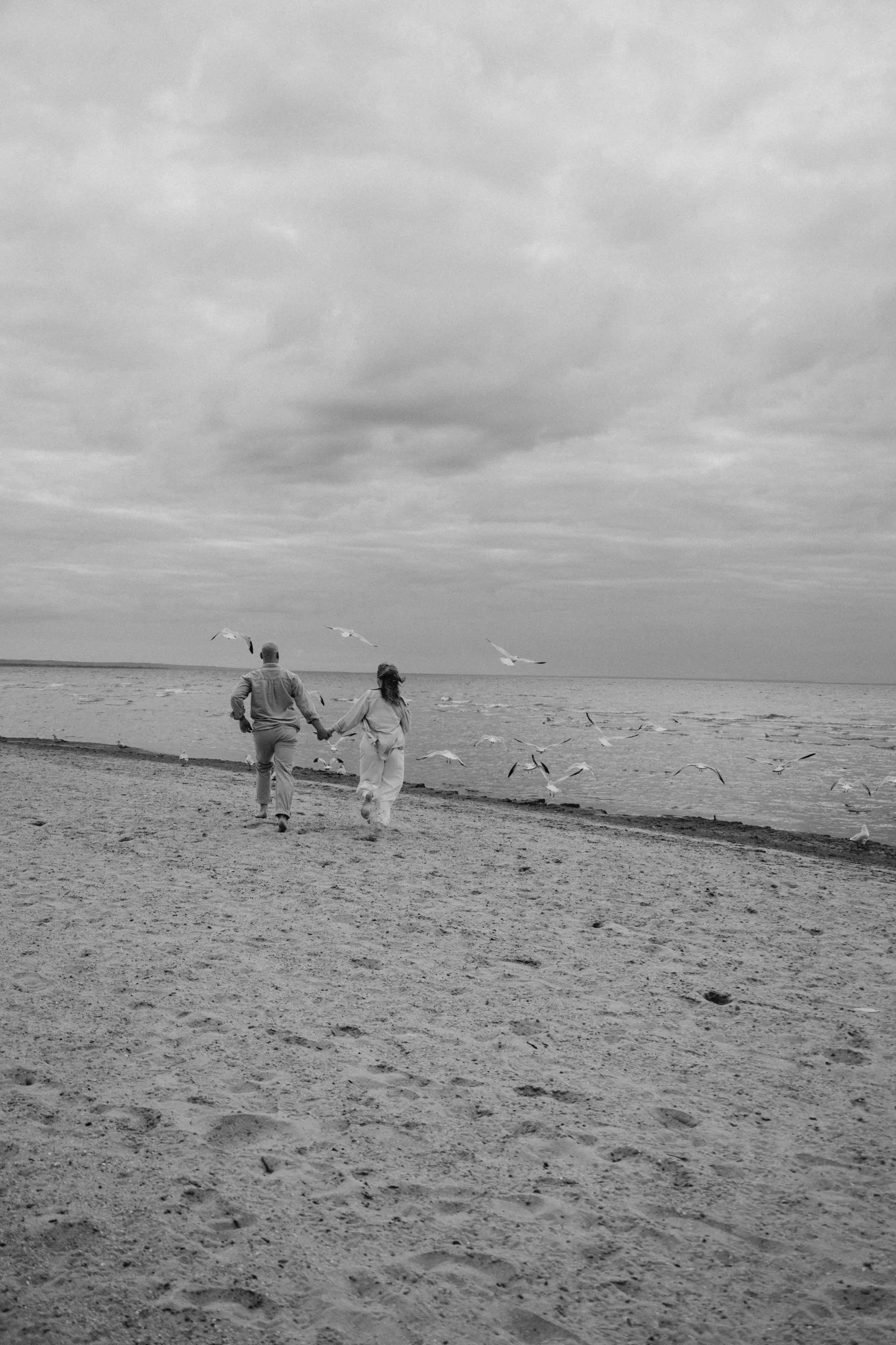 A black and white photo of a couple walking hand in hand along a sandy beach near the ocean, with seagulls flying overhead and cloudy sky.
