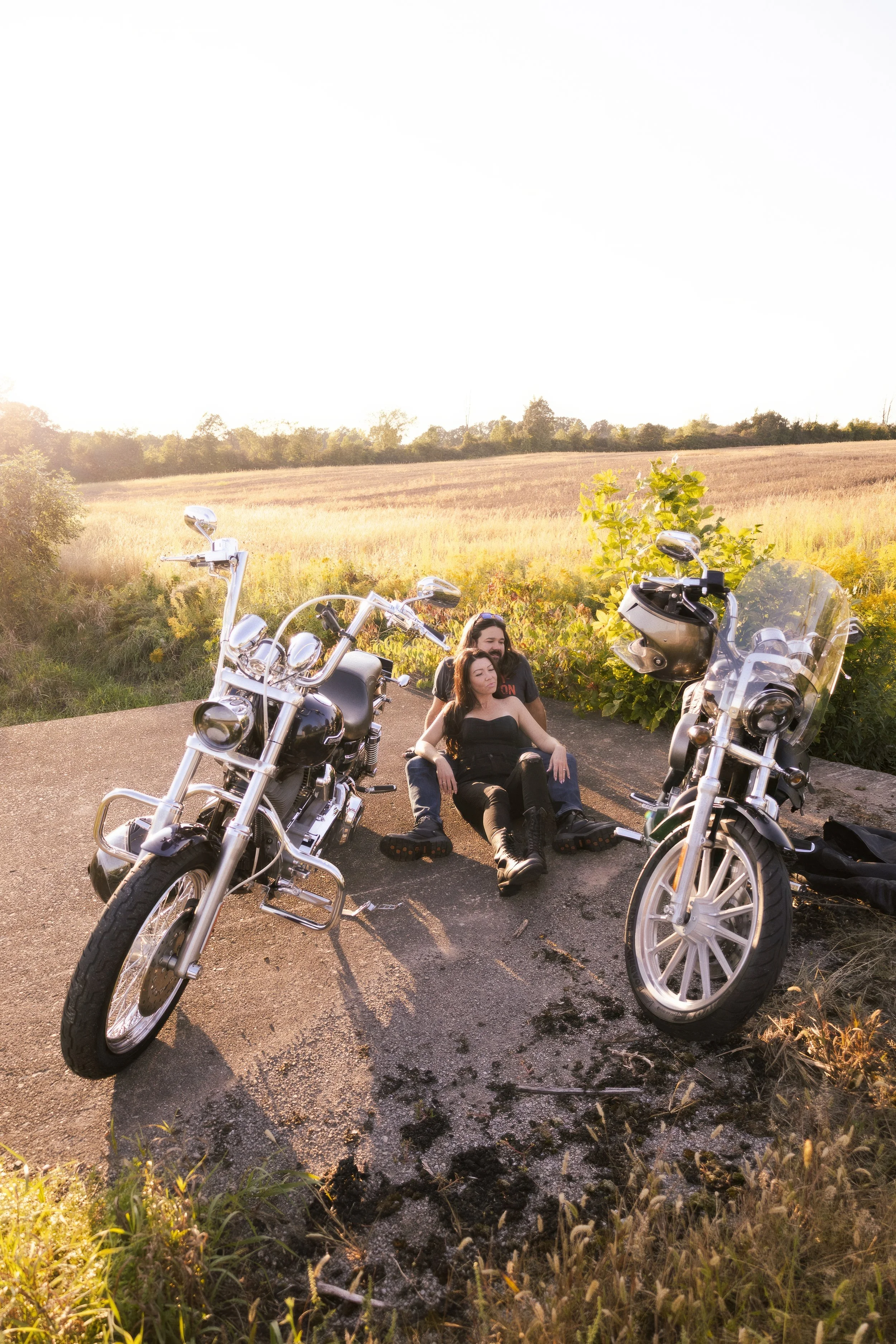 A man and woman sitting on the ground between two parked motorcycles in a rural setting during sunset.