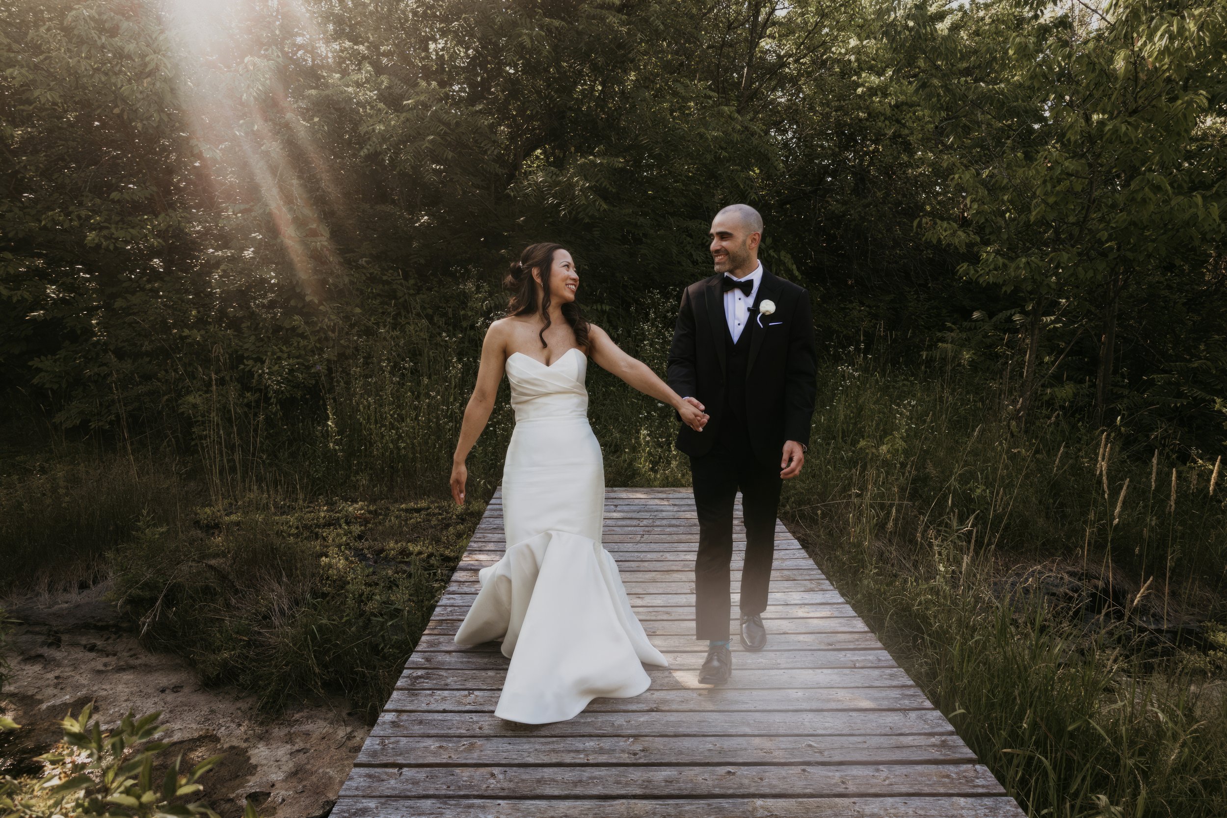 A bride and groom walking hand in hand on a wooden bridge through a wooded area, smiling at each other, during their wedding.
