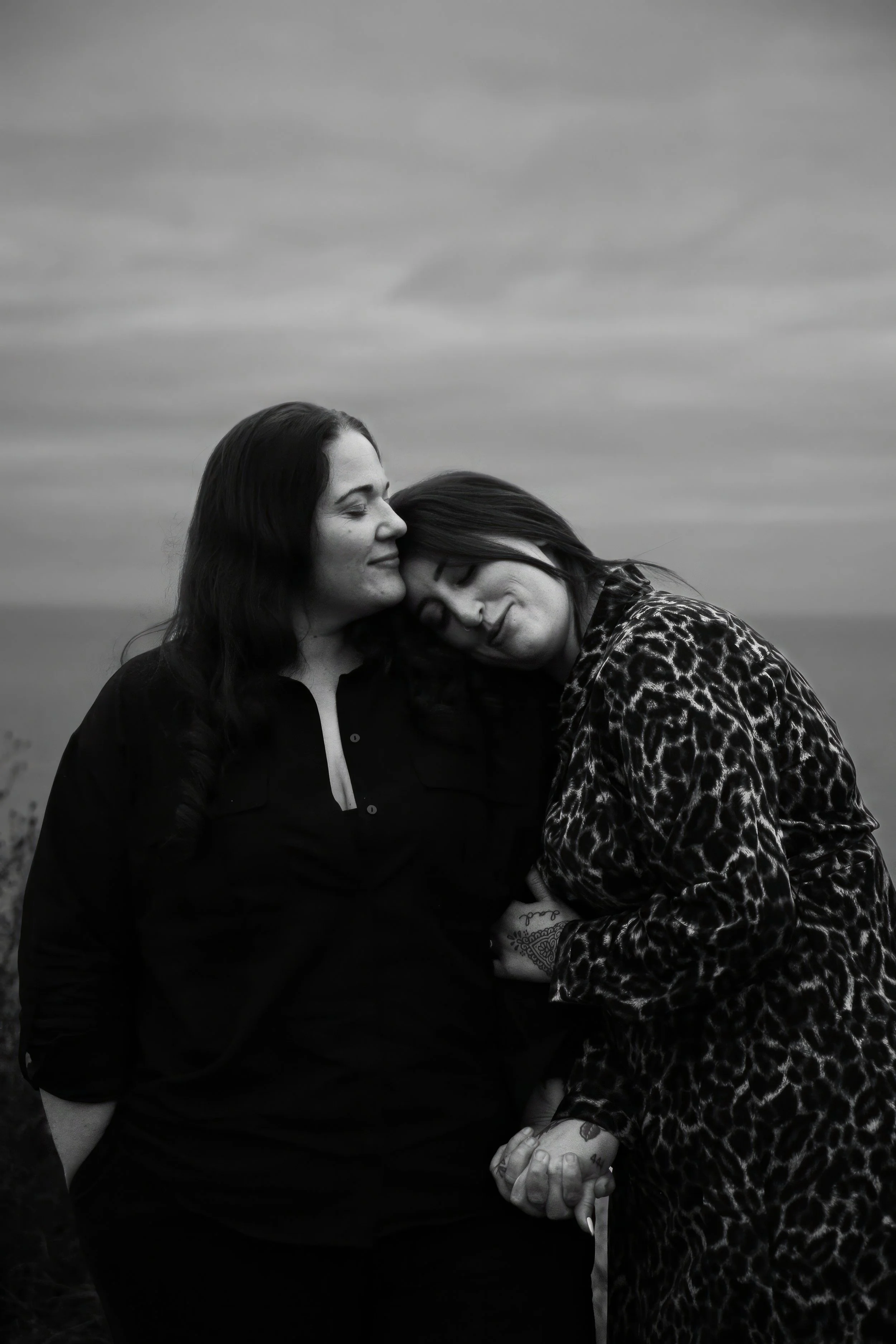 Two women sharing an intimate moment outdoors, with their eyes closed and faces touching, one wearing a dark shirt and the other wearing a leopard print jacket, holding hands.