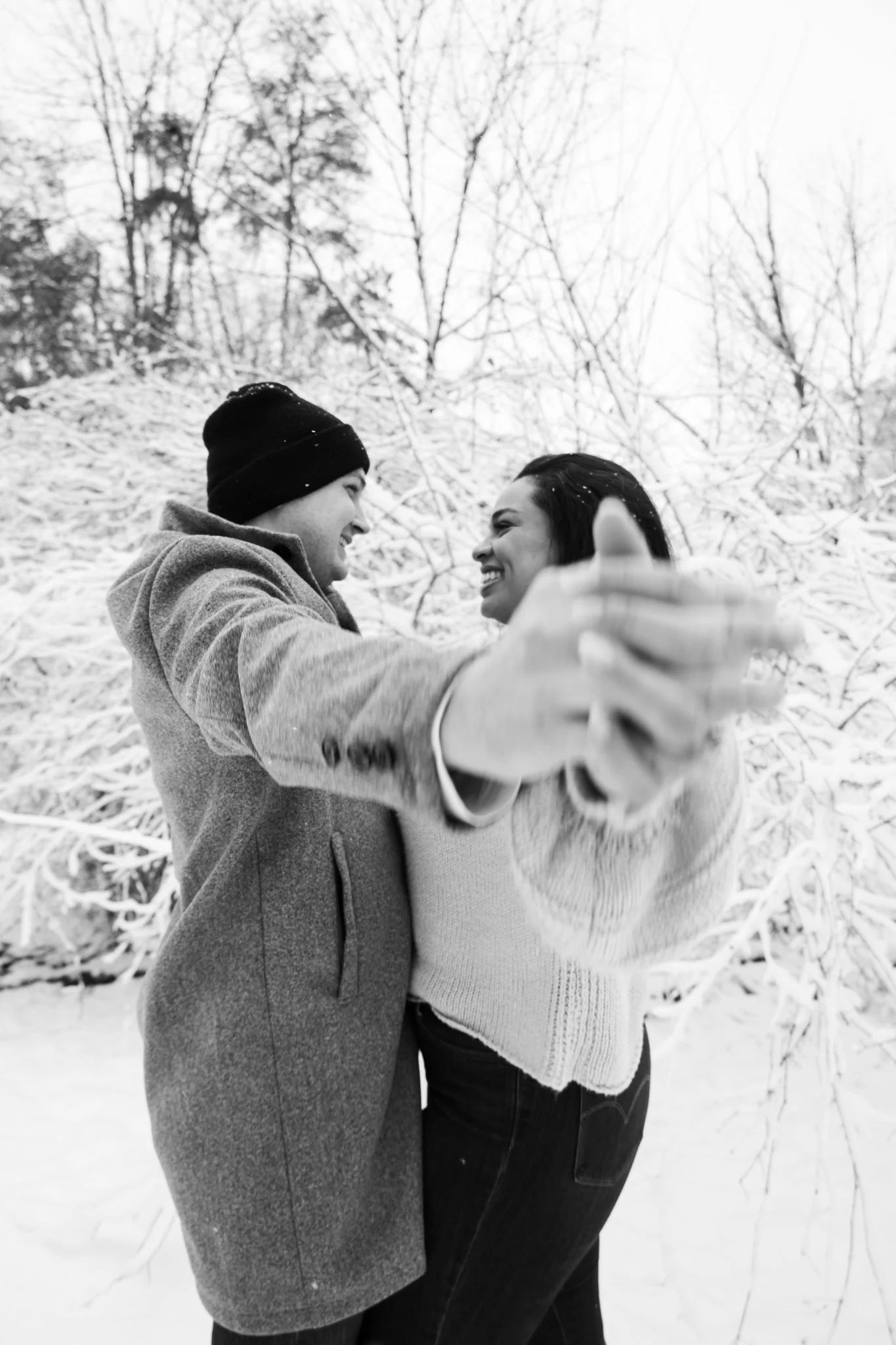 A couple embracing outdoors in snowy weather, smiling and holding hands, with snow-covered trees in the background.