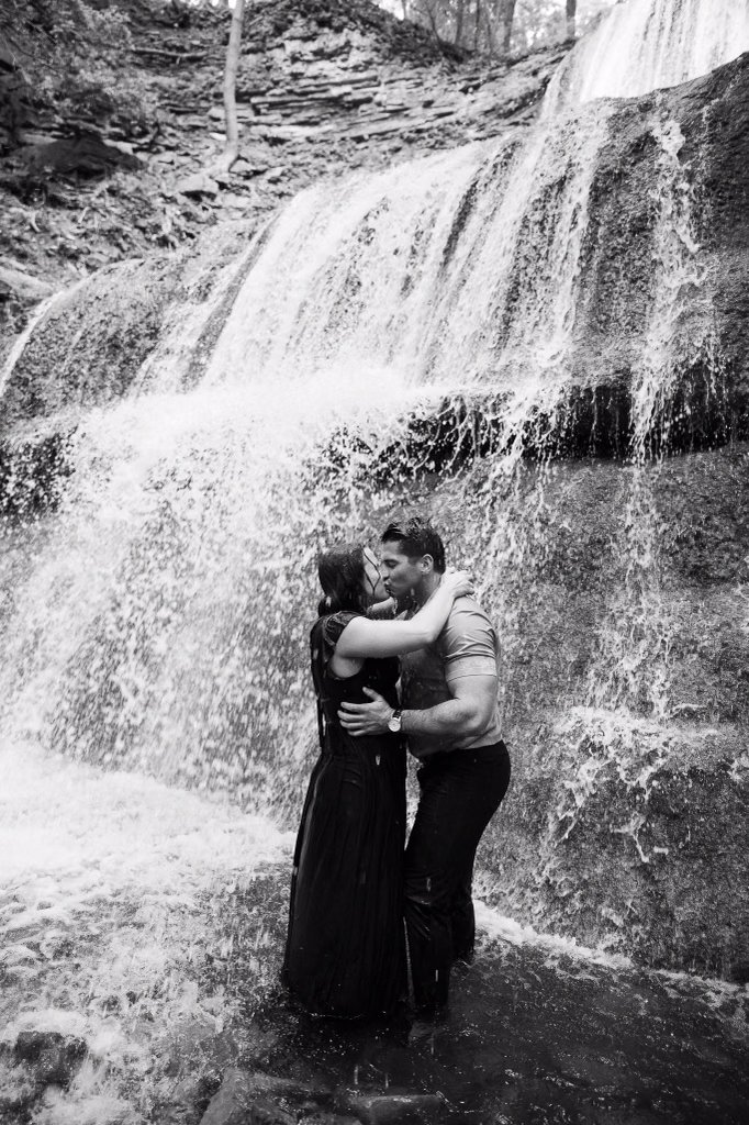 A couple kissing under a waterfall, standing in the water, captured in black and white.