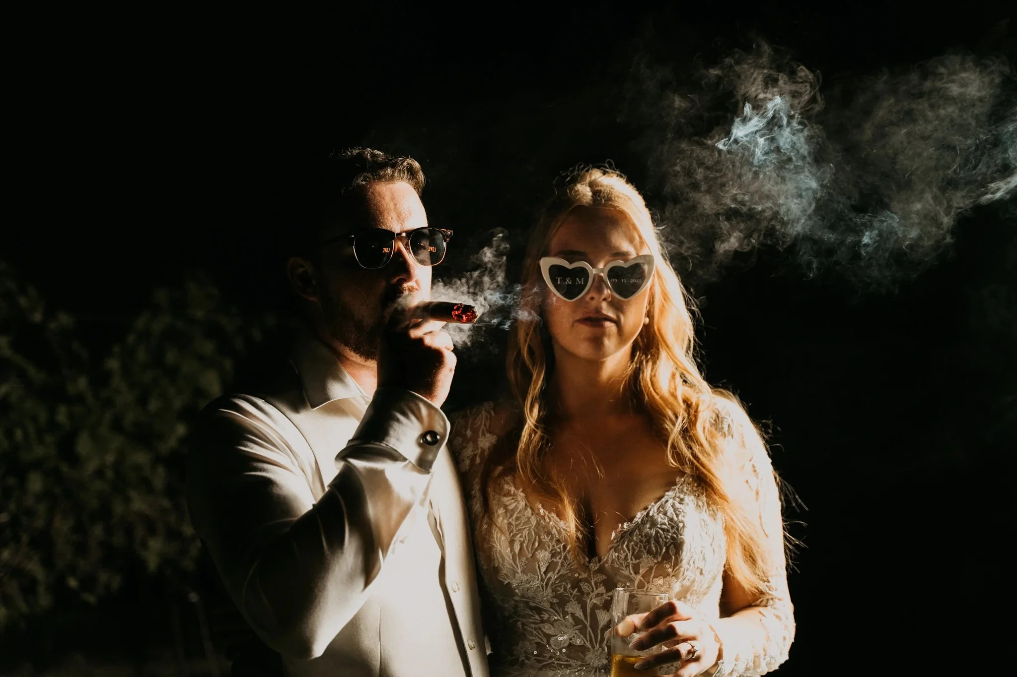 A man in a tuxedo and sunglasses is smoking a cigar, standing next to a woman in a lace dress and heart-shaped sunglasses at night with smoke around them.