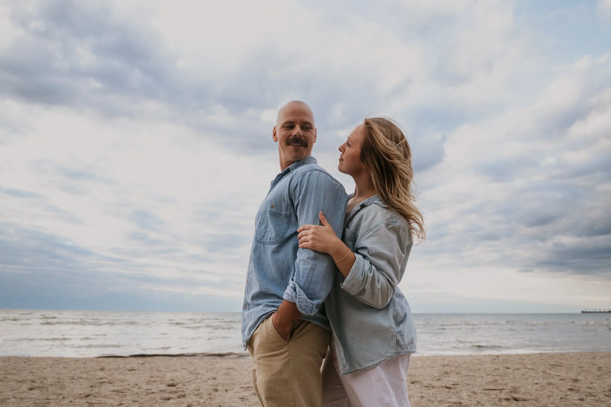 A couple stands close together on a beach, with the ocean and cloudy sky in the background. They are facing each other, with the woman holding the man's arm. Both are dressed in casual denim jackets, and the man has a mustache and is smiling.