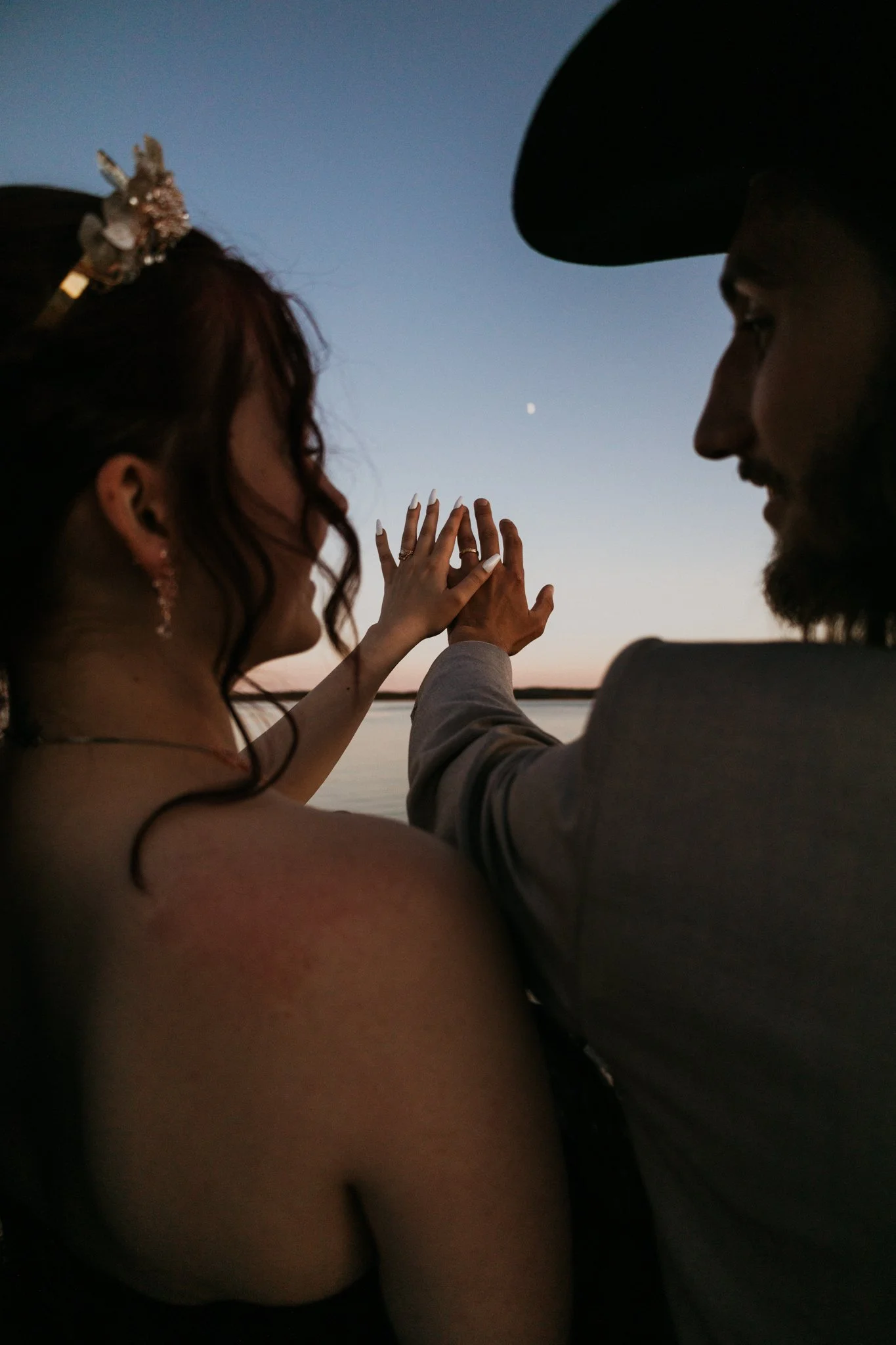 A couple touching hands during sunset by a body of water, with the woman wearing a bridal veil and earrings, and the man wearing a cowboy hat.