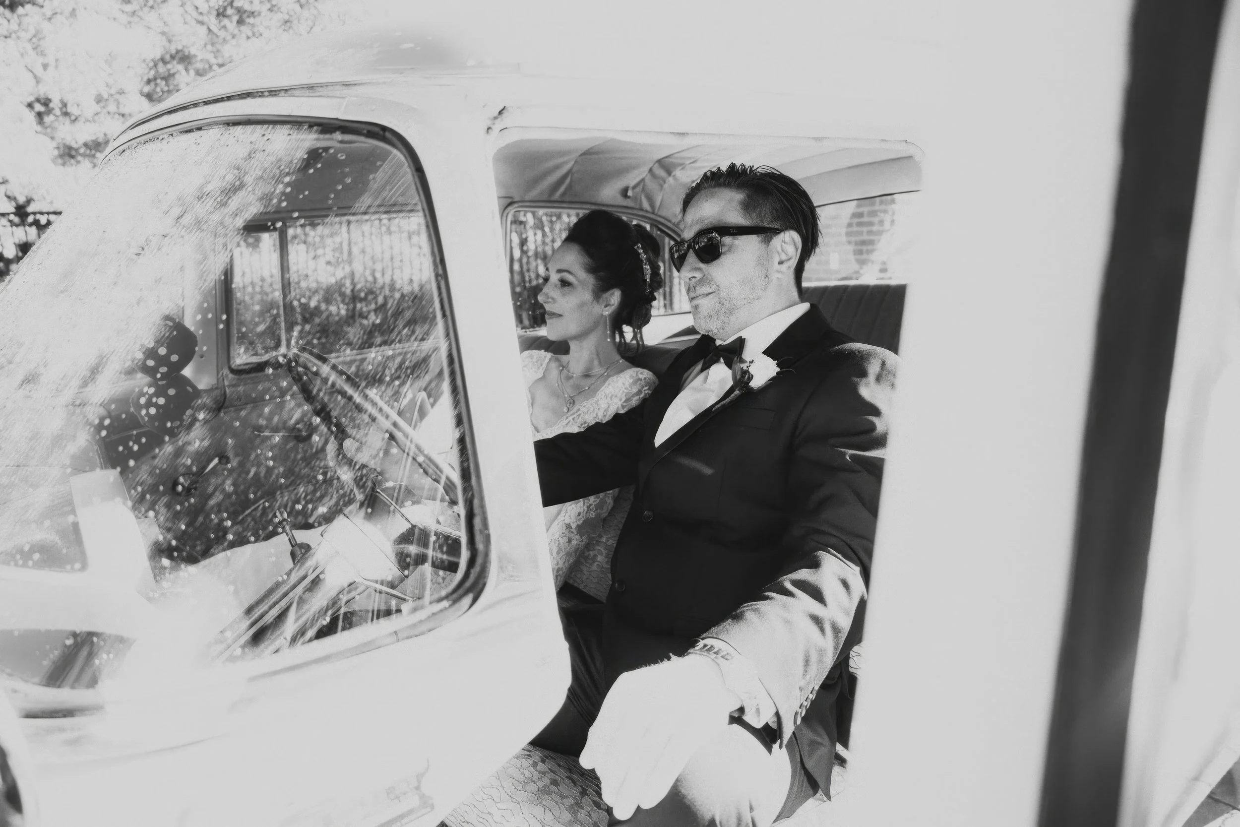 Black and white photo of a bride and groom sitting in the front seat of a vintage car, dressed in wedding attire, with the bride wearing a lace dress and the groom in a tuxedo and sunglasses.