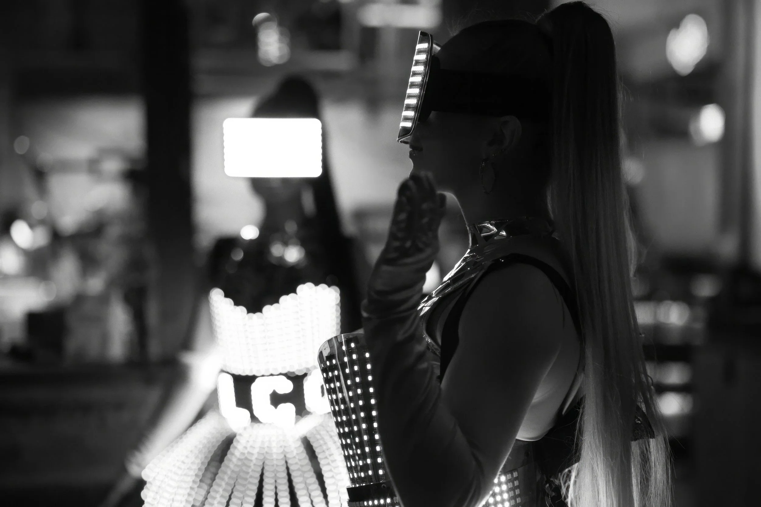 A woman in a futuristic costume with LED lights on her arm and a headpiece, standing in profile against a brightly lit background, possibly at a nightclub or event.