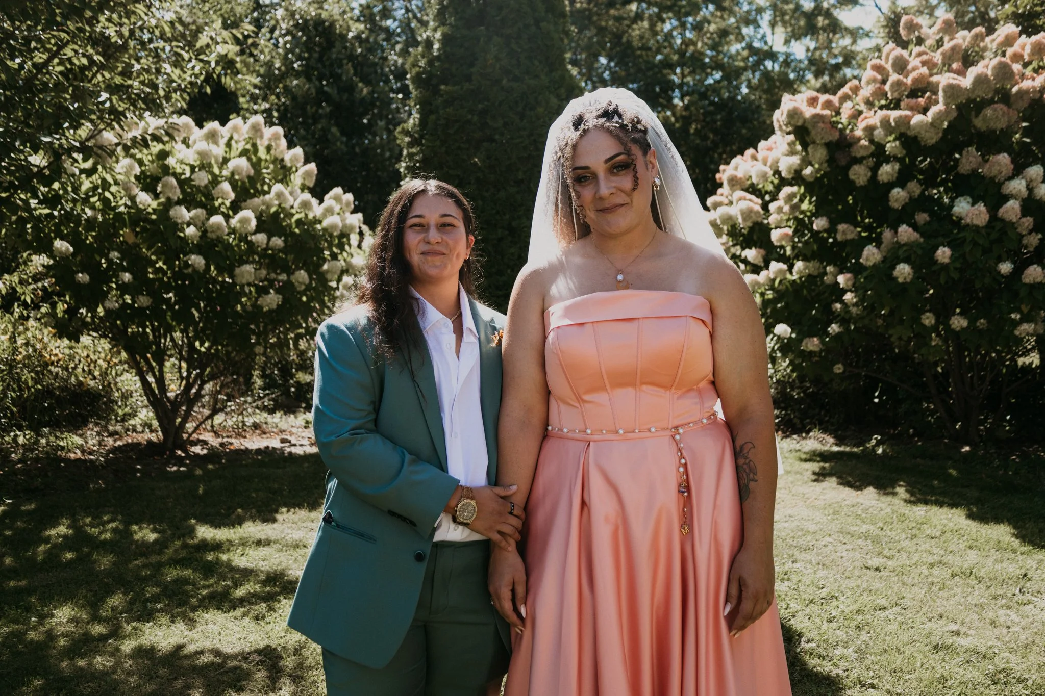 Two women standing outdoors during daytime with flowering bushes in the background. One woman is in a green suit and white shirt, and the other is in a peach-colored dress with a veil, earrings, and necklace.