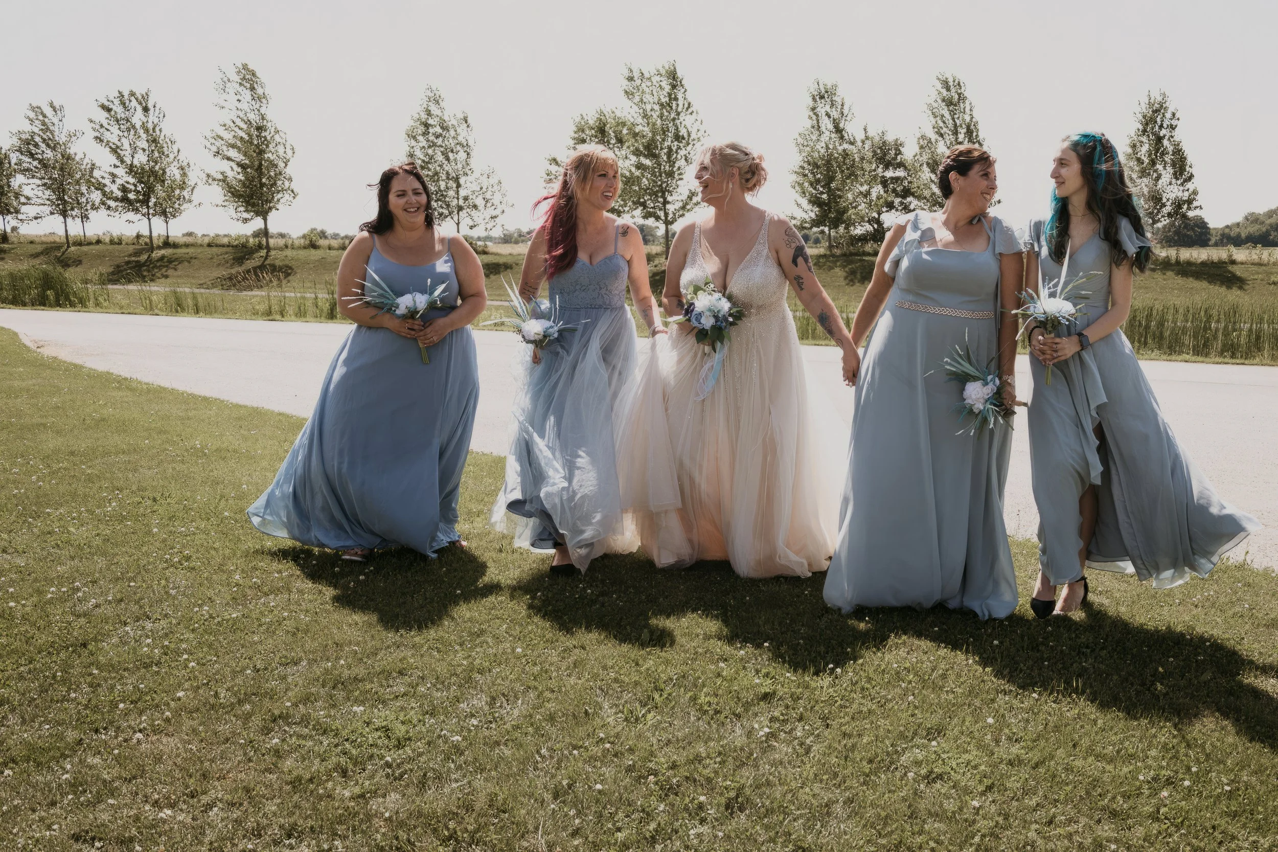 Group of six women in pastel dresses, walking outdoors holding bouquets on a sunny day