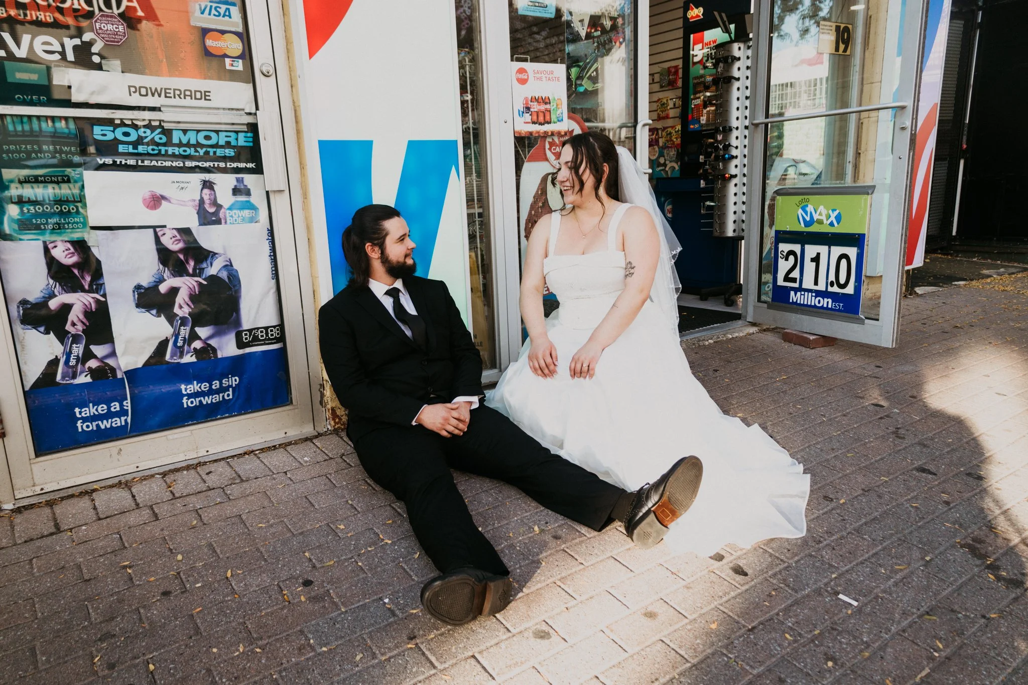 A man in a black suit and a woman in a white wedding dress sit on the sidewalk outside a store, smiling and looking at each other. The woman is standing and the man is sitting.