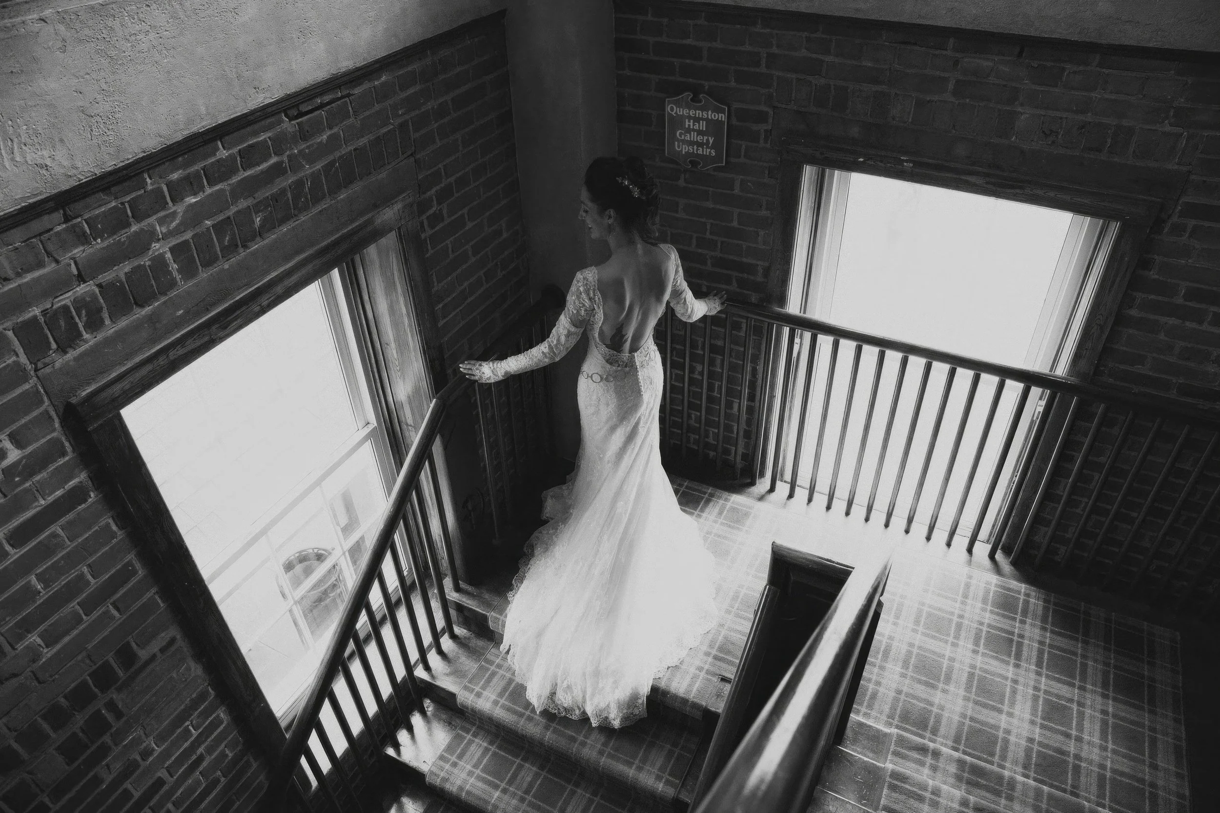 A woman in a wedding dress descending a stairway near large windows, with brick walls and a sign that reads 'Queenston Hall Gallery Upstairs'.