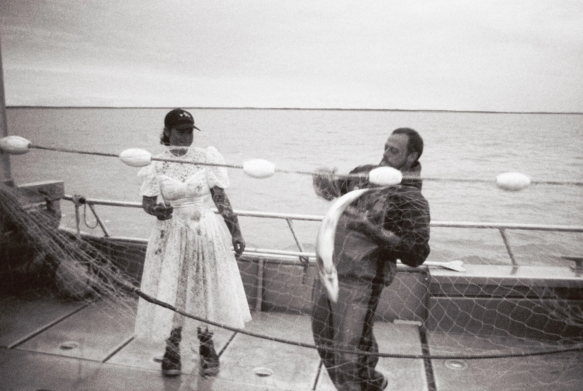 A woman and man standing on a boat deck by the water, with fishing nets and buoys, during the day.