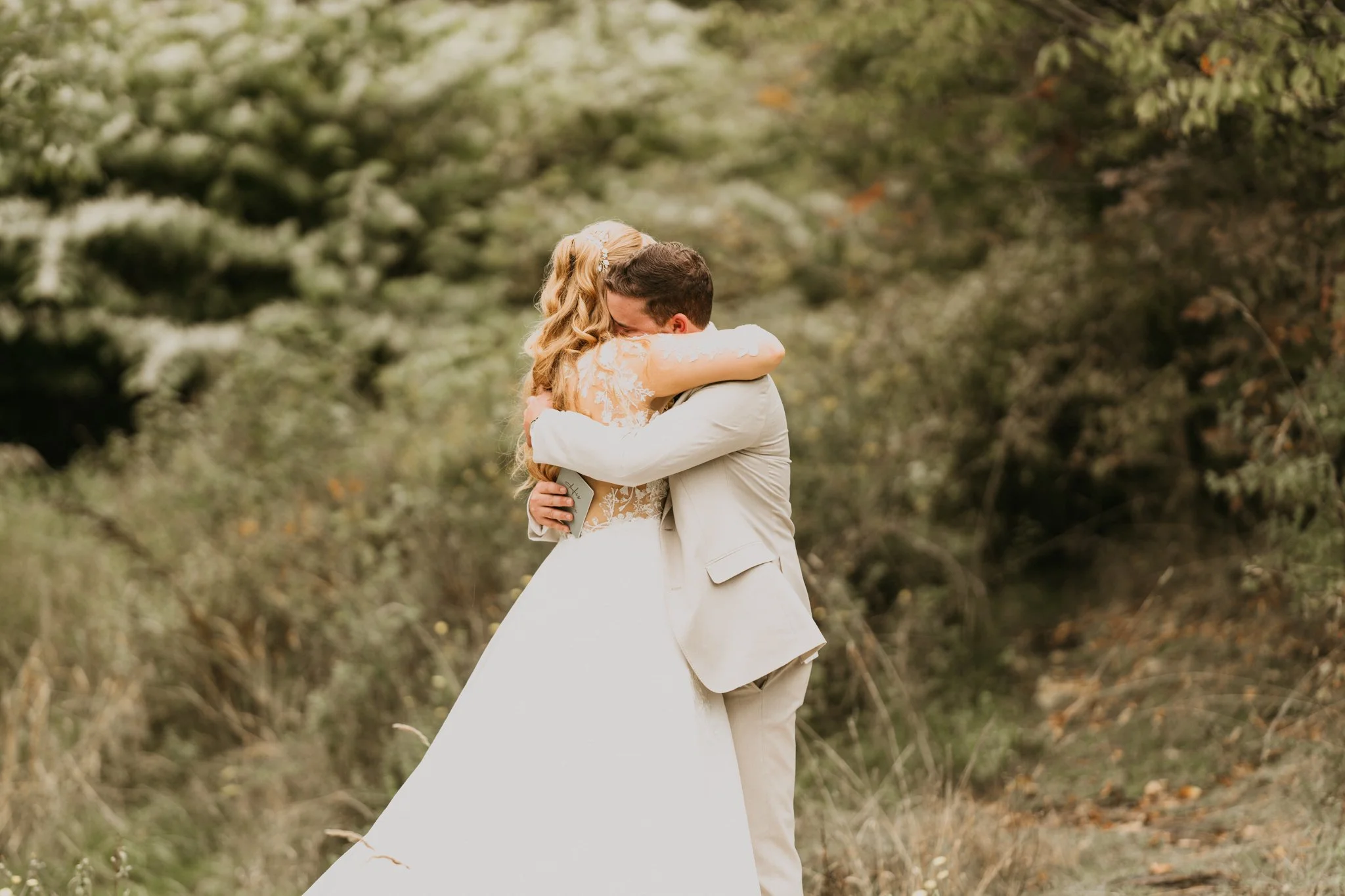 A bride and groom hugging in an outdoor setting with trees and foliage in the background.
