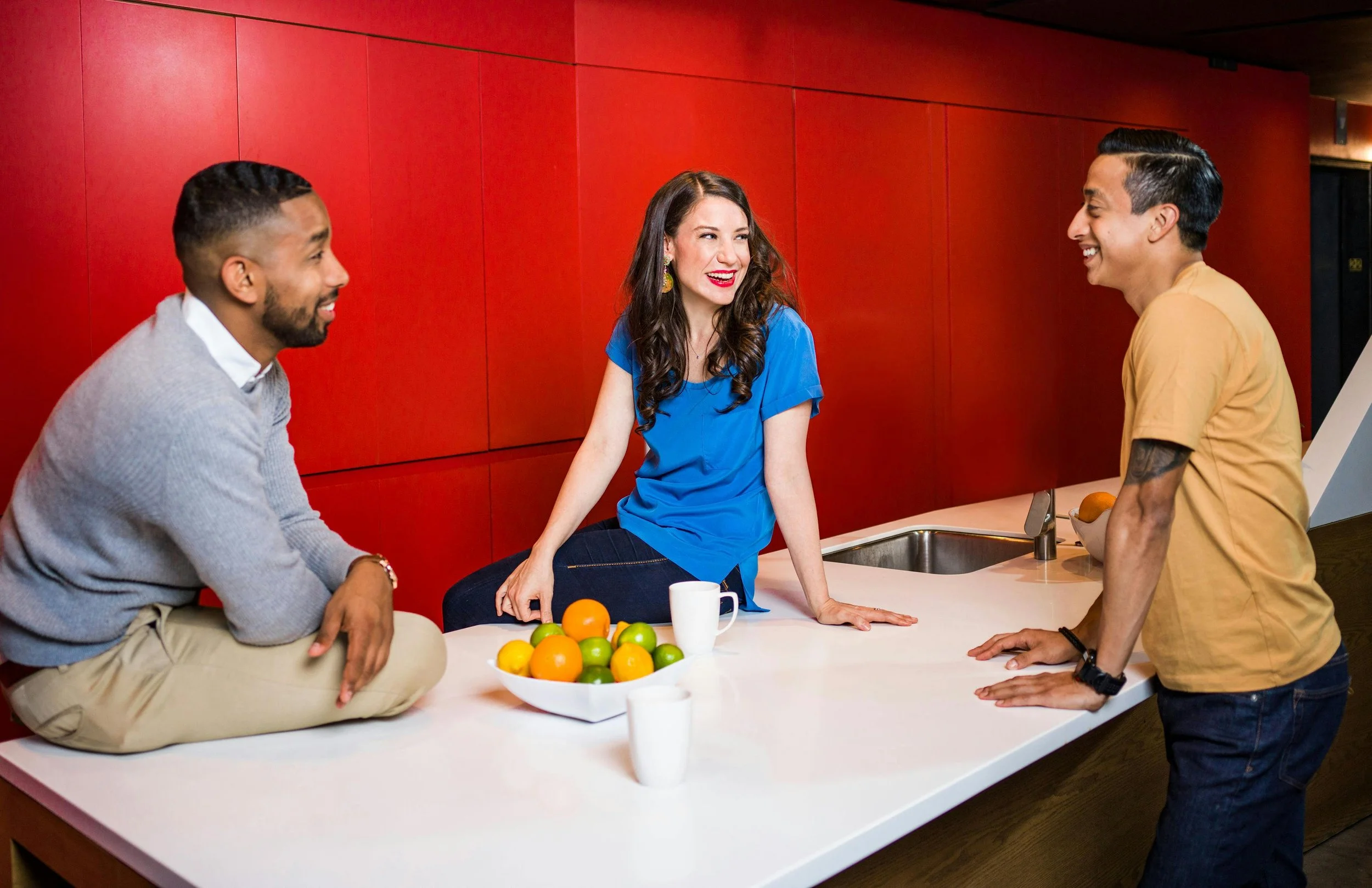 Three gender-diverse and racially-diverse people sit and stand around a counter, looking and smiling at each other, with a red wall behind them.